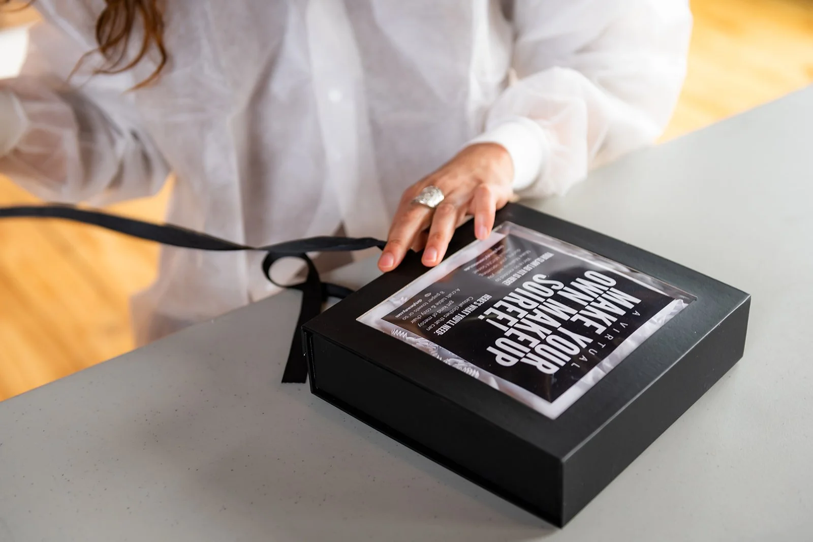 A person wearing a white shirt opening a black box with a transparent cover that has white text and a ribbon handle, on a gray table.
