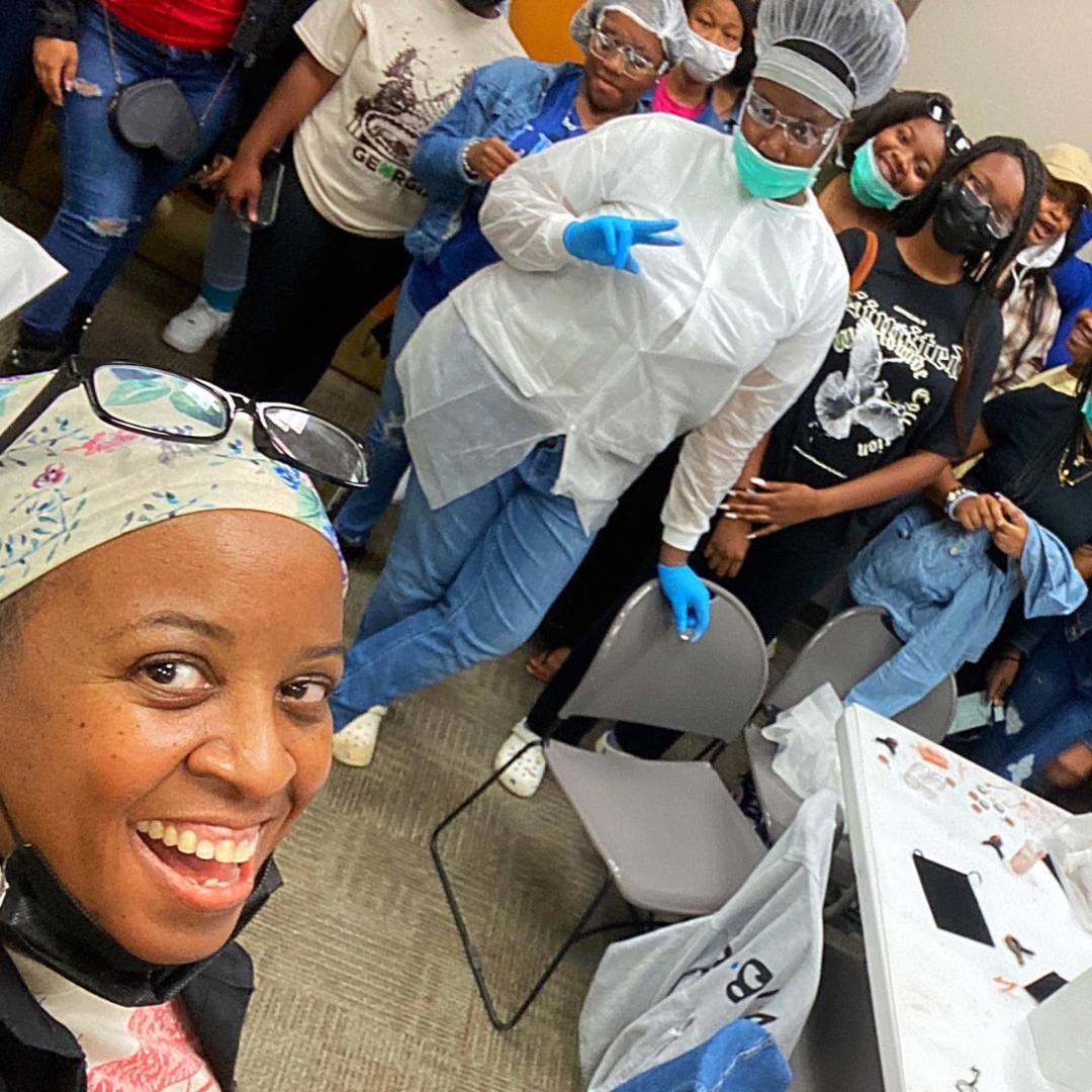 A woman taking a selfie in a room filled with people, some wearing masks, gloves, and protective gowns, with a table nearby covered with items and papers.