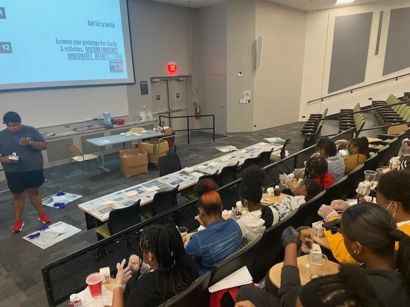 A classroom lecture hall with students sitting in tiered seats, some wearing masks, and a person standing at the front near a table with supplies, a large screen displaying instructions.