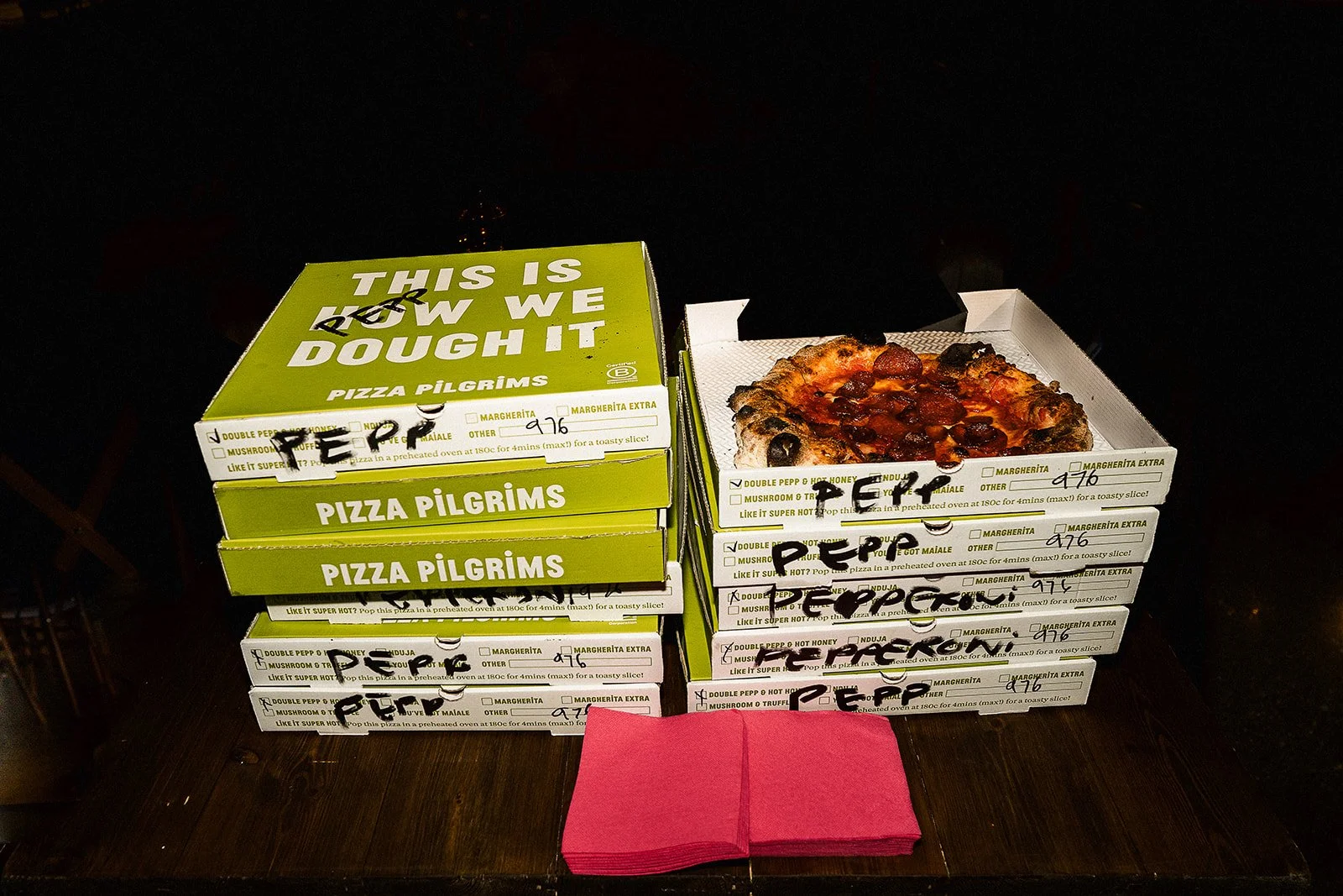 Multiple pizza boxes labeled "Pizza Pilgrims" stacked on a table, one box open showing a slice of pepperoni pizza, red napkins in front.