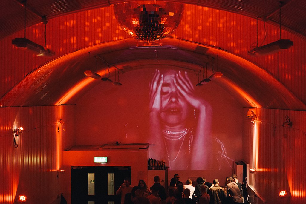 People watching a large projection of a woman crying with her hand on her forehead in a dimly lit, red-colored theater or event space with a curved ceiling and disco ball.