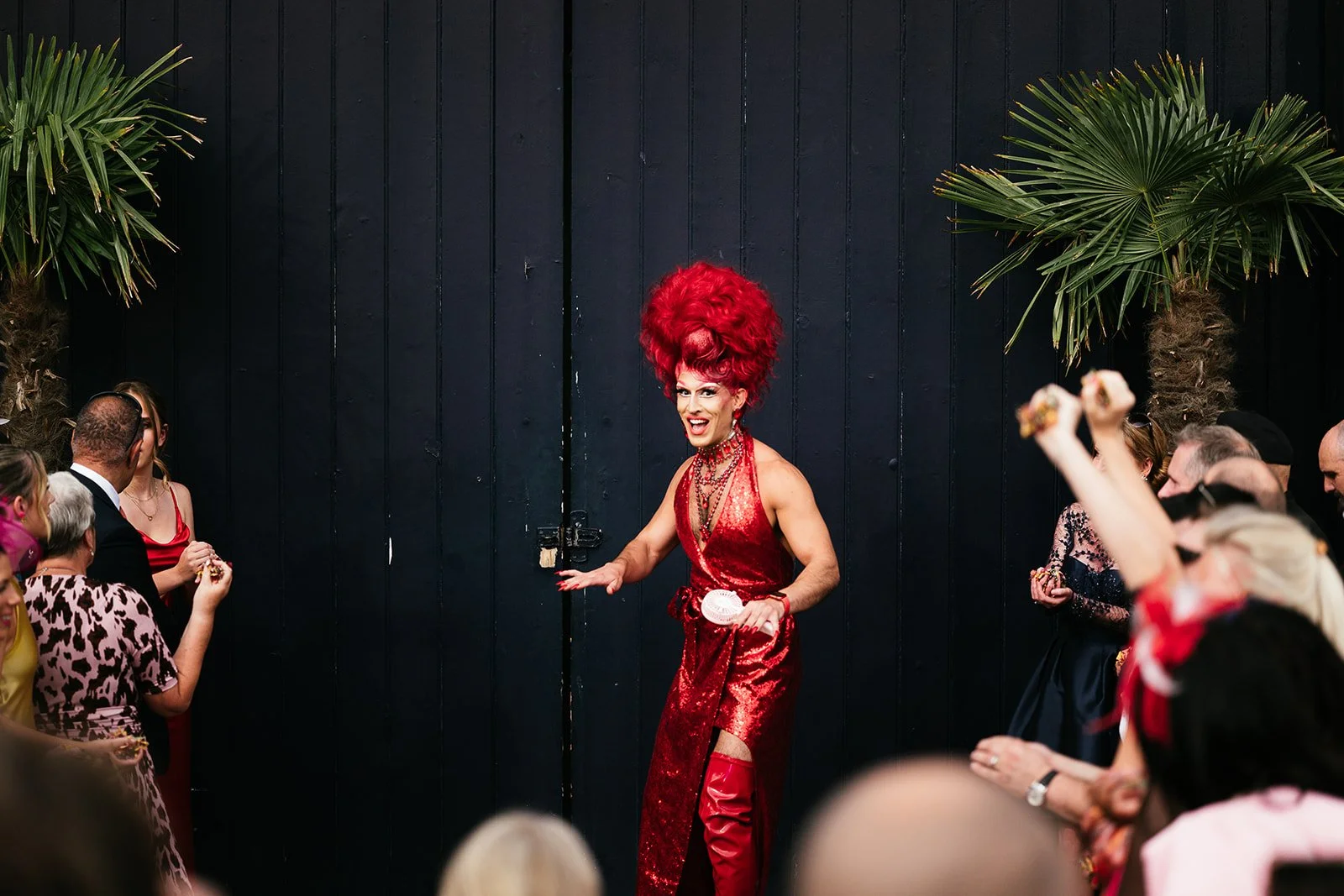 A drag queen with bright red hair and a shiny red dress performing in front of an audience at an indoor event, framed by palm trees on either side.