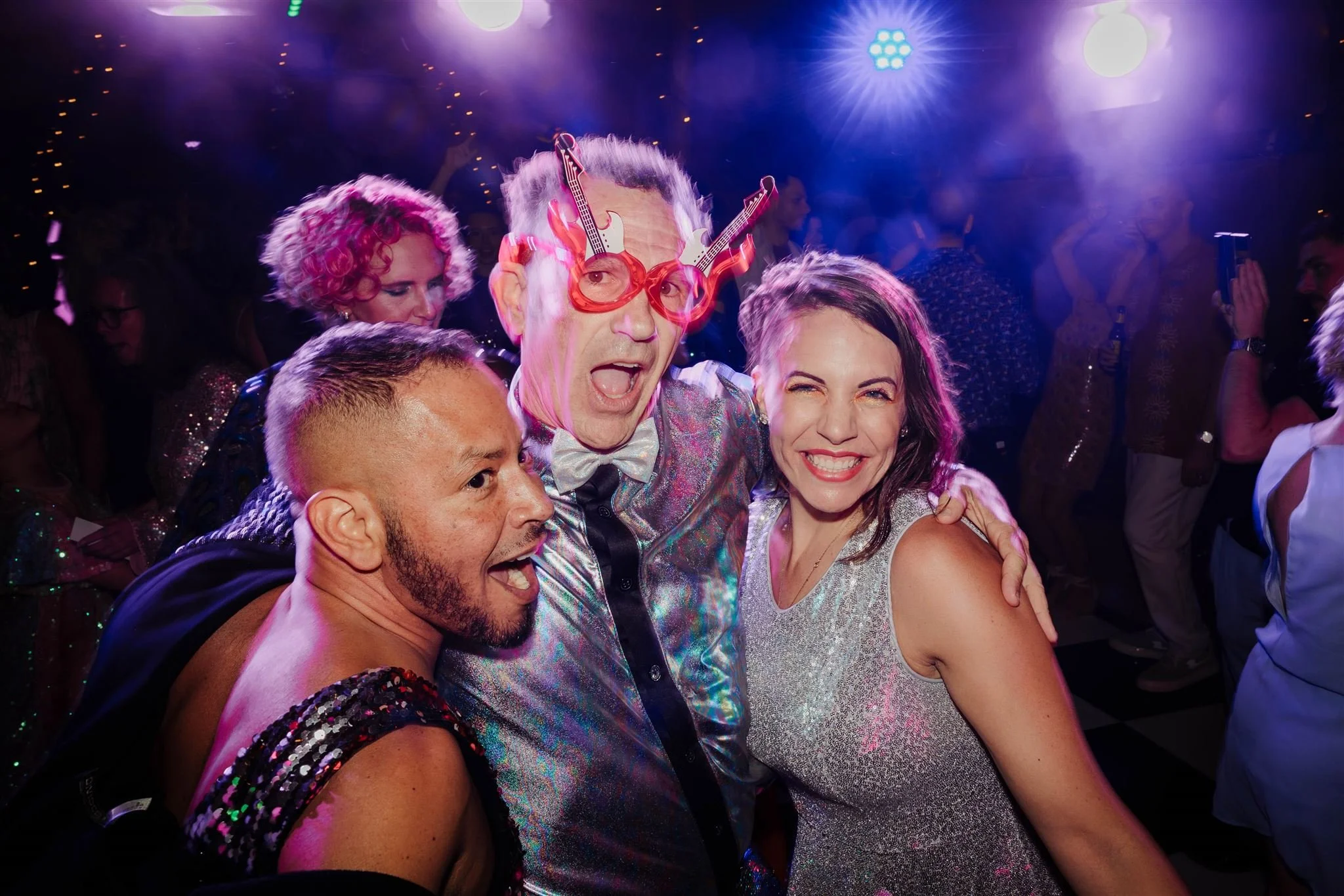 People celebrating at a party, smiling and having fun with colorful lighting and festive attire.