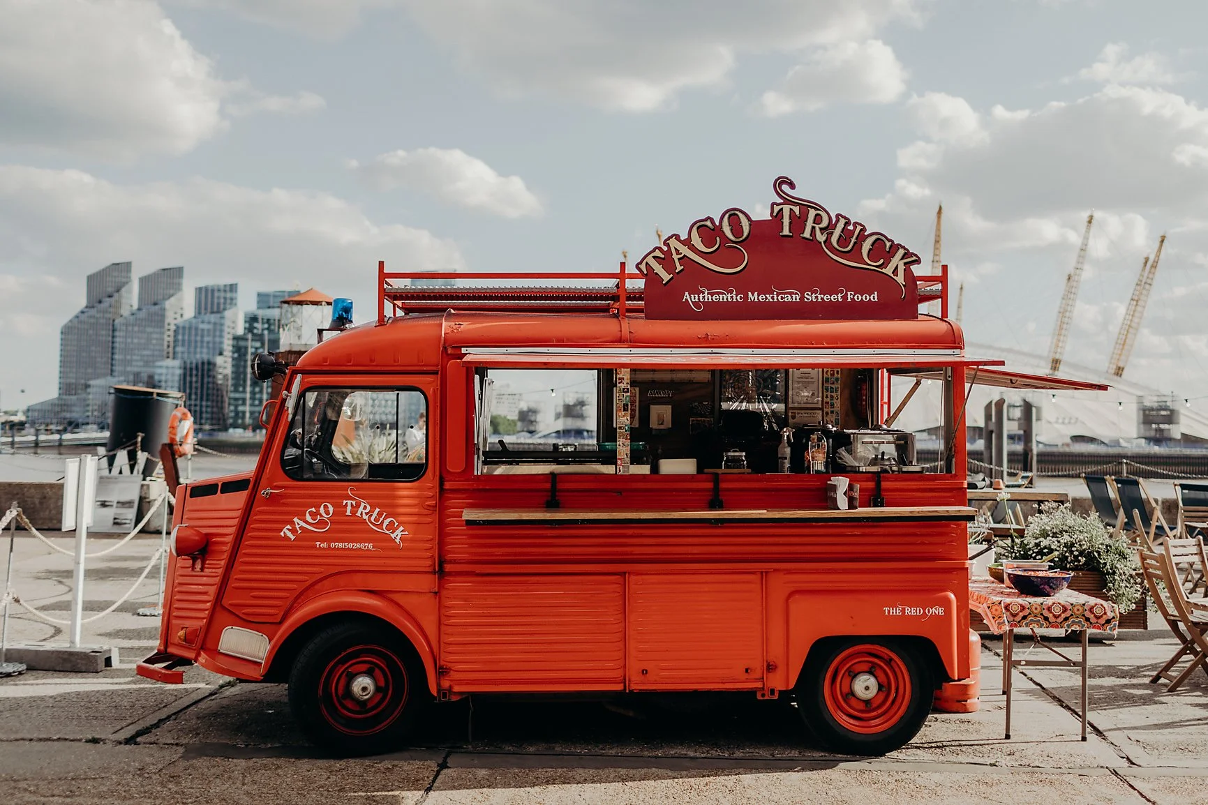 A red taco truck labeled 'Taco Truck' with a sign that says 'Authentic Mexican Street Food' on a street with clouds in the sky.