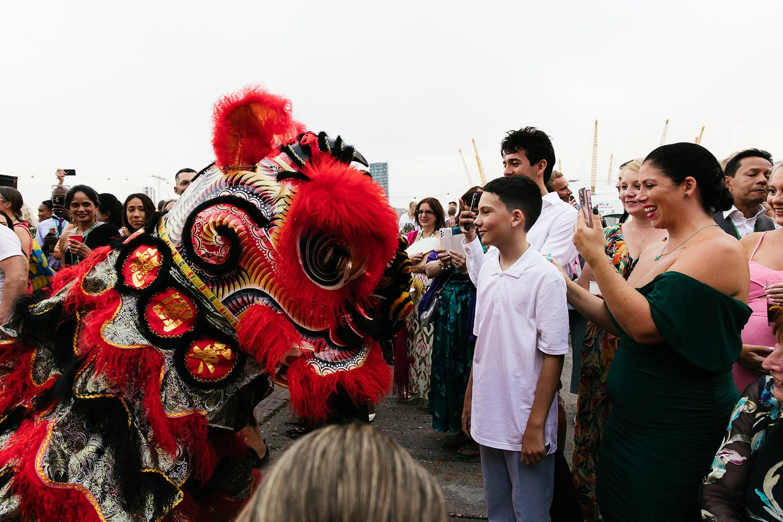 People gathered outdoors watching a colorful lion dance performance, taking photos and smiling.