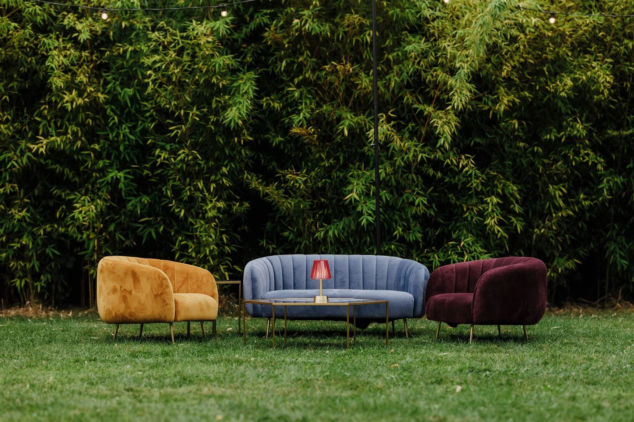 Outdoor seating arrangement on grass with a blue velvet sofa, a yellow velvet armchair, a burgundy velvet armchair, a glass-top coffee table, and a small red table lamp, with dense green foliage in the background.