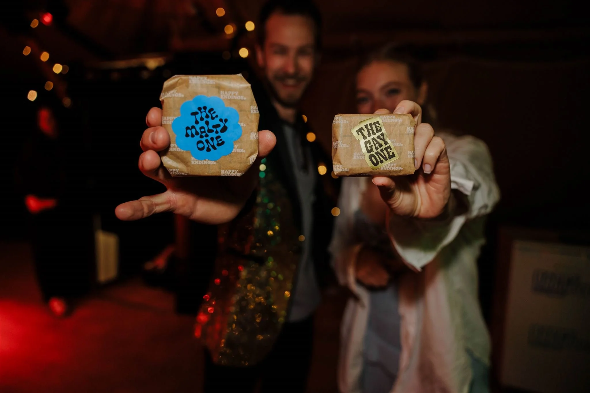 Two people holding wrapped gift boxes labeled "The Male One" and "The Gay One" in a dimly lit room decorated with holiday lights.