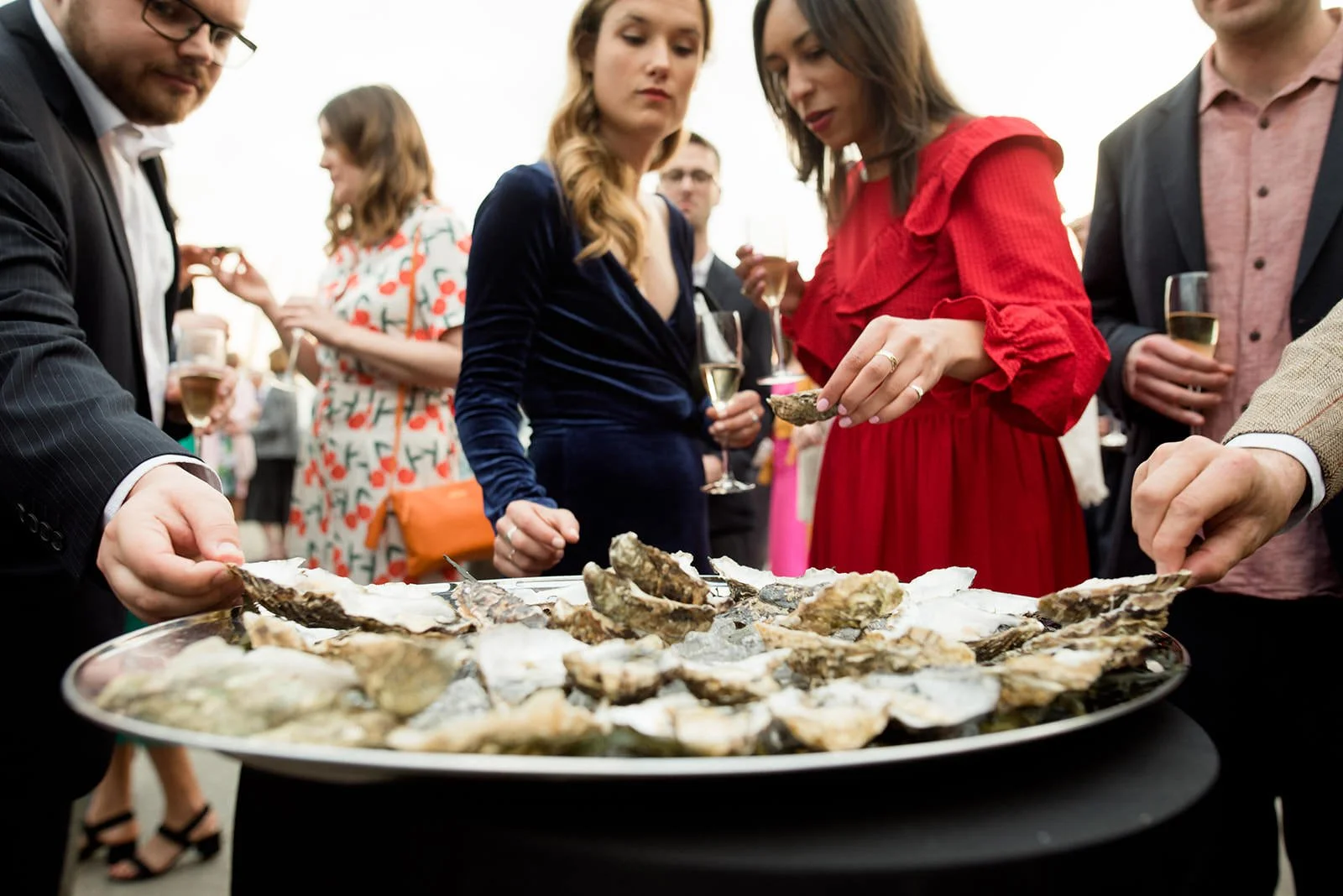 People at a social gathering serving and eating oysters, some holding glasses of champagne, dressed in semi-formal attire.