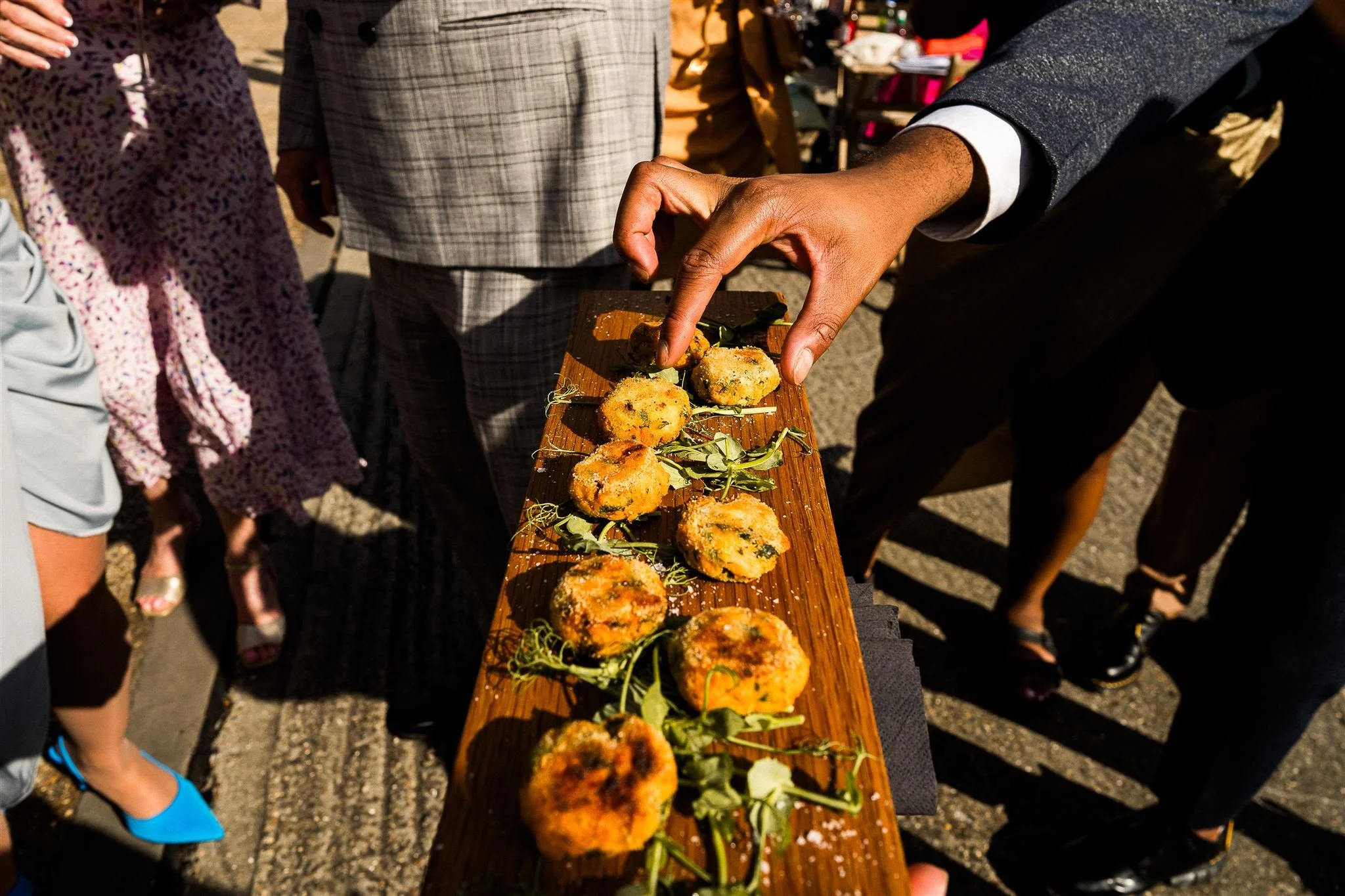 Person serving breaded appetizers on a wooden board during an outdoor event, with people dressed in business attire and casual clothing nearby.