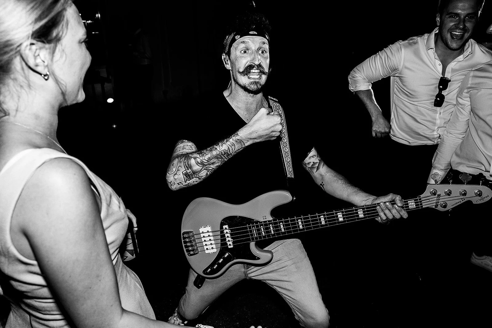 Black and white photo of a man playing an electric bass guitar, surrounded by smiling people at a lively social gathering.
