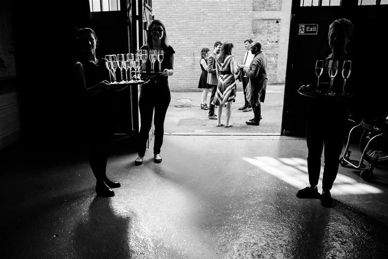 Three women holding trays of champagne glasses inside a venue and a group of people outside in conversation.