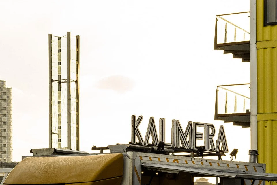 Street view showing a building with a KALIMERA sign on the roof, adjacent to a tall modern building with glass windows and a yellow building with balconies.