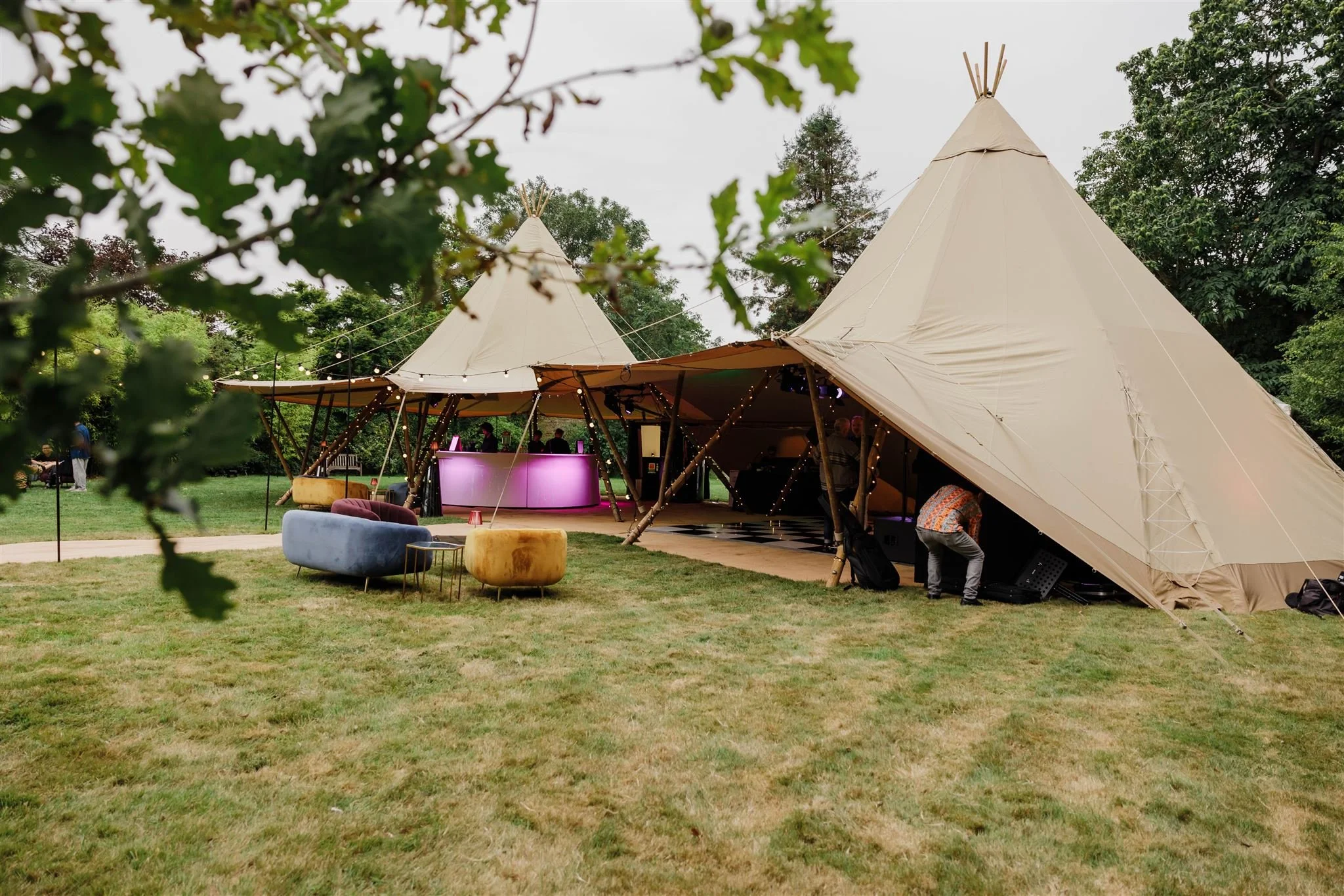 A large outdoor event tent with a dance floor, surrounded by green trees and grass, with colorful seating area in front.