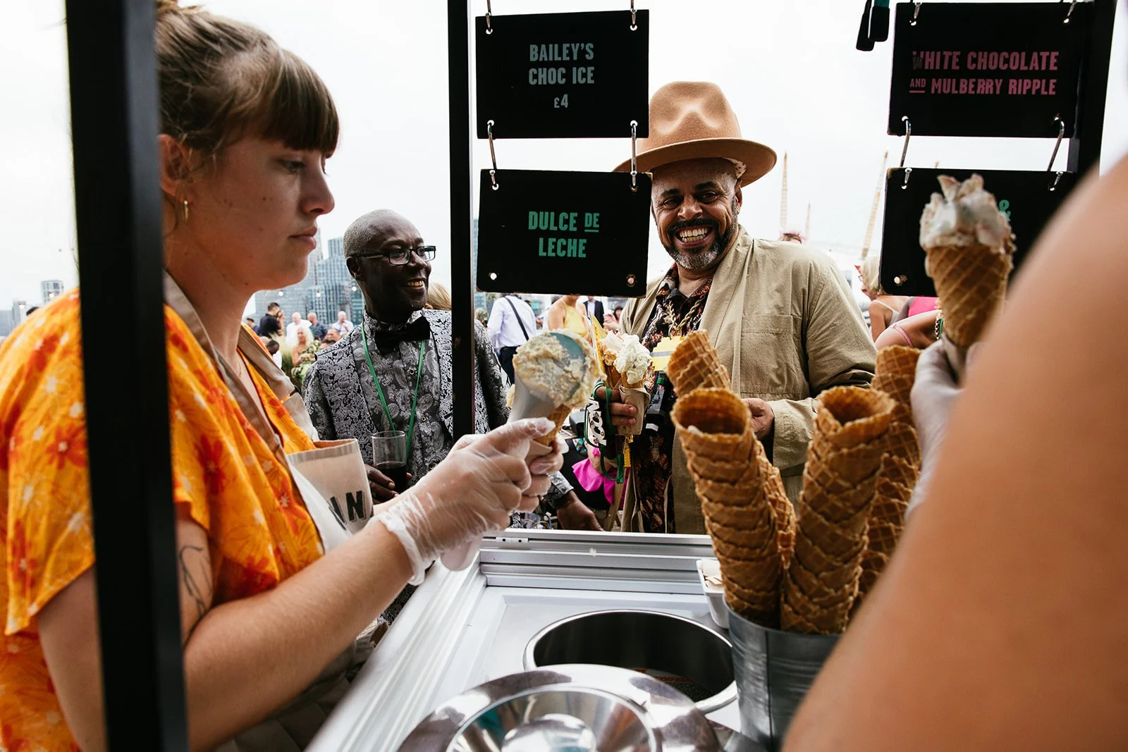 People at an outdoor ice cream stand with signs listing flavors such as Bailey's Choc Ice and Dulce de Leche, serving ice cream cones to smiling customers.