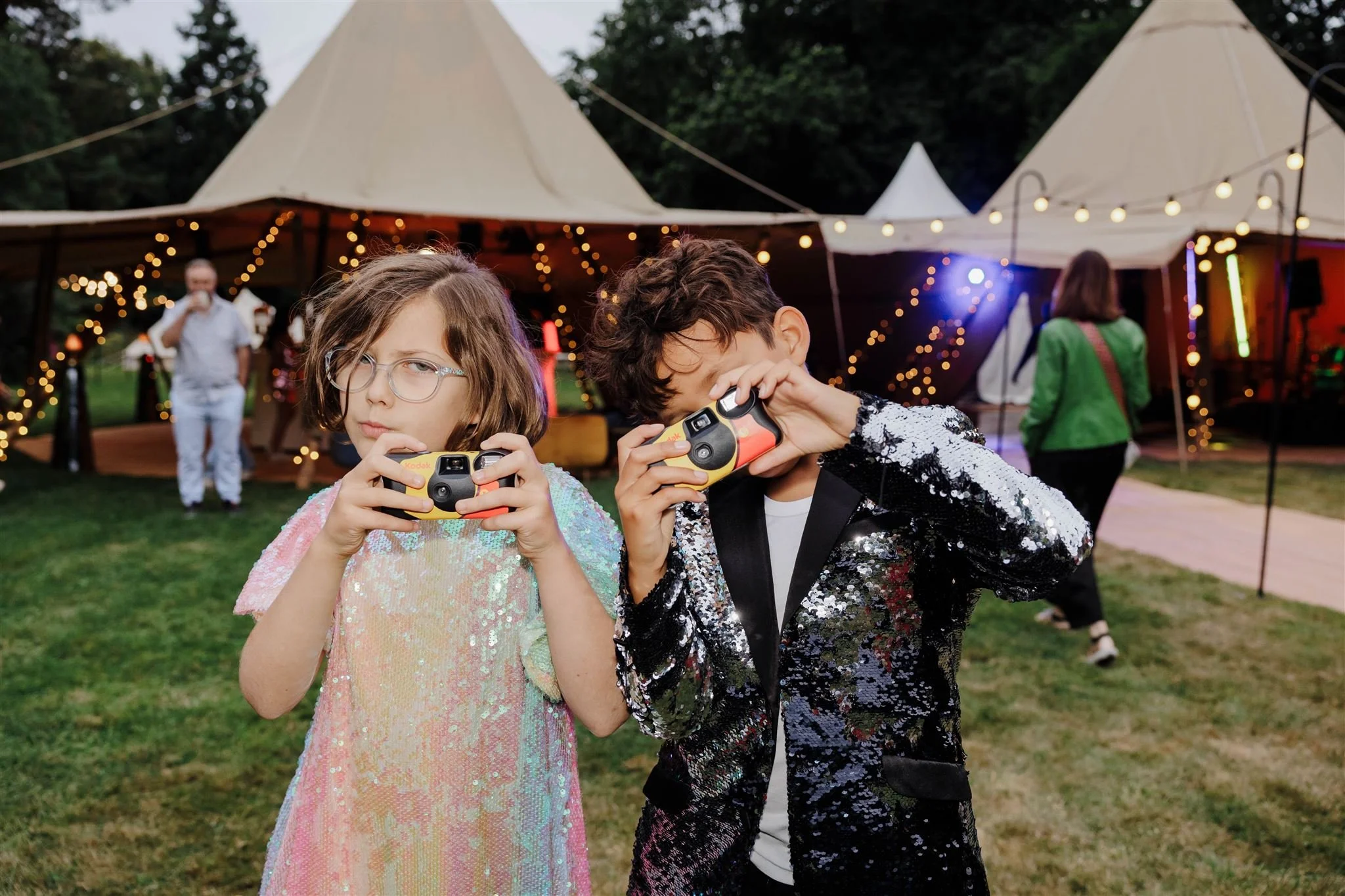 Two children with cameras at a festive outdoor party with tents, festoon lights, and other guests in the background.