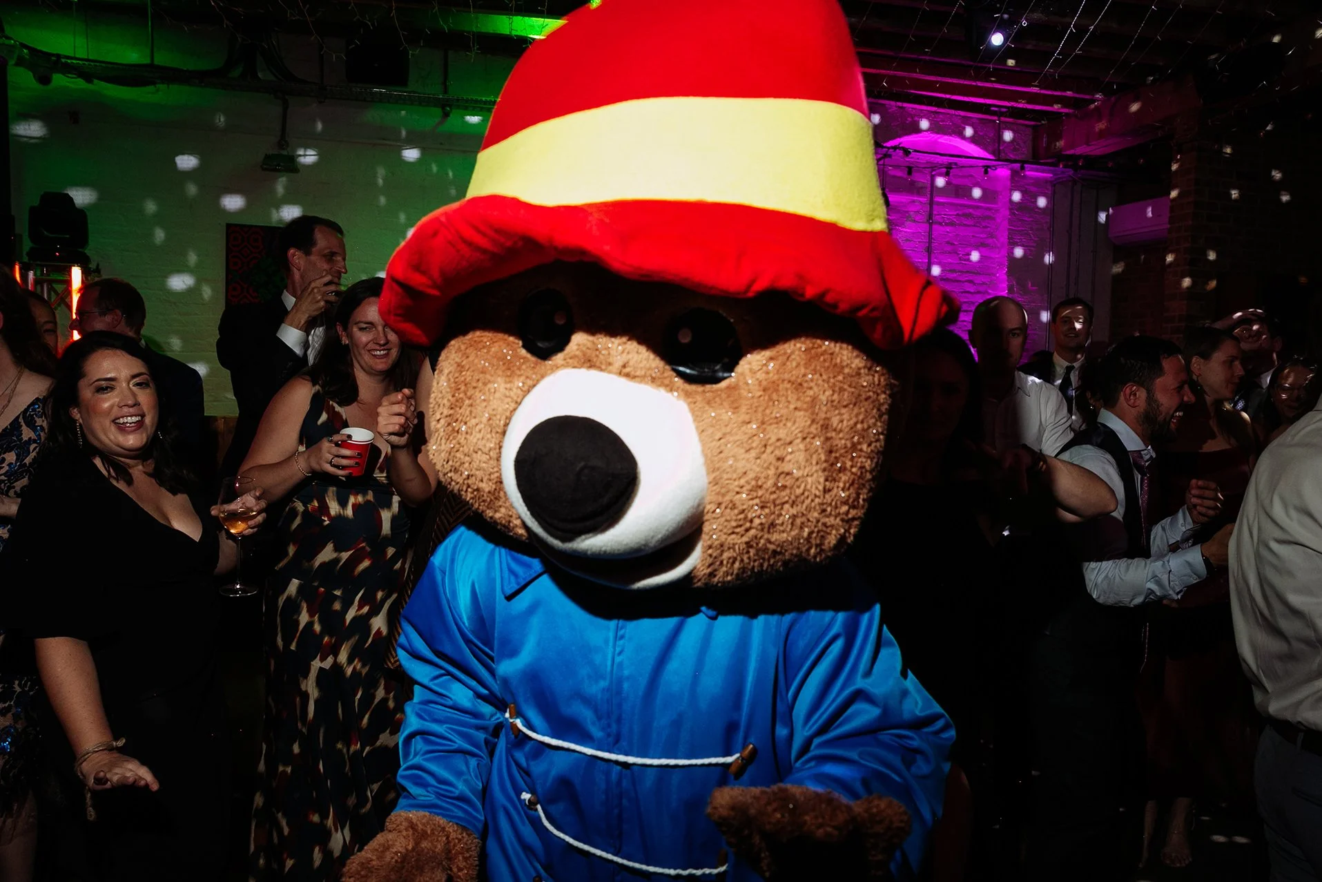 A person in a bear costume wearing a red and yellow hat and a blue jacket at a lively party with people dancing and smiling.