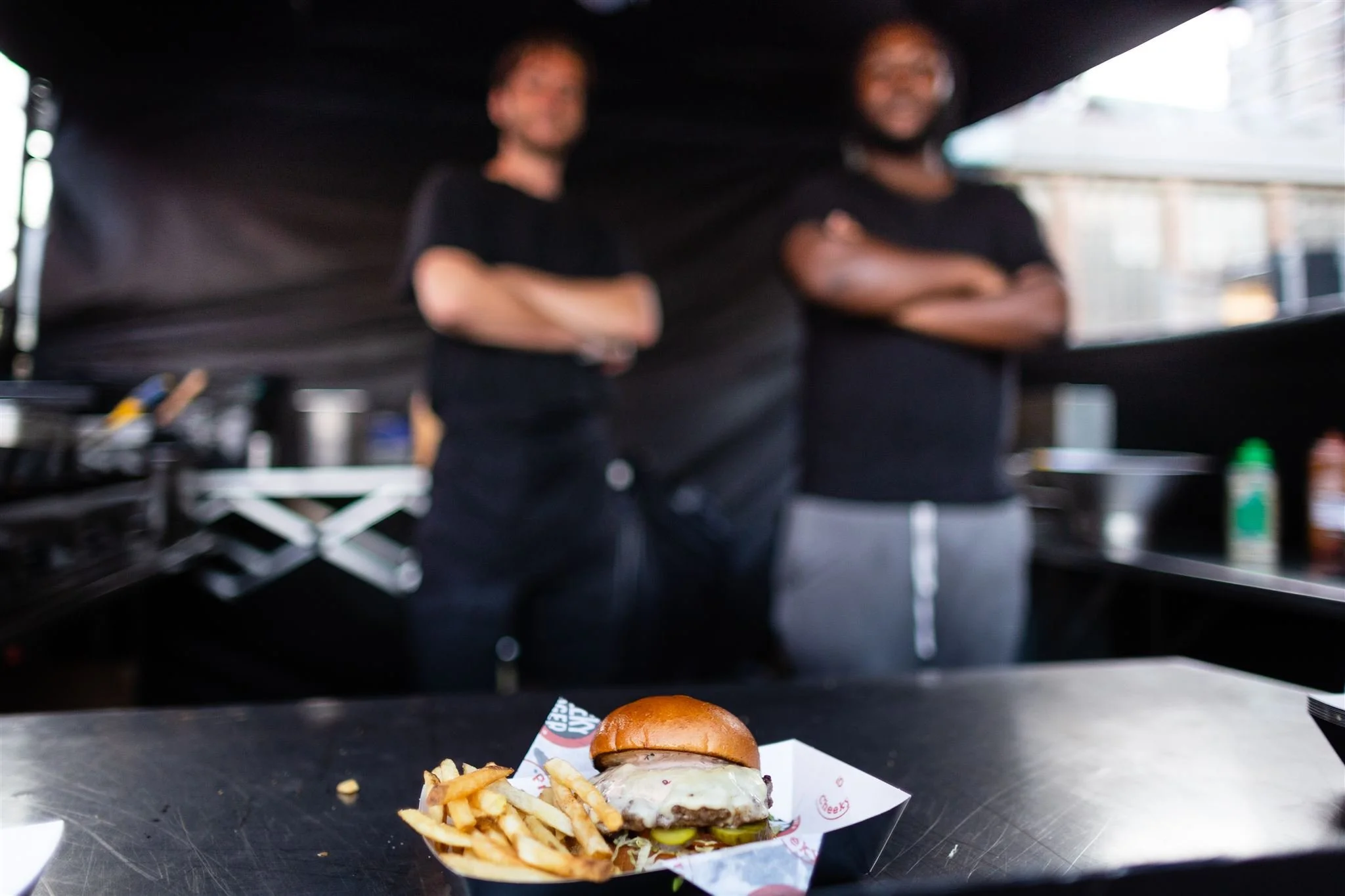 A cheeseburger with lettuce, pickles, and cheese served with fries on a paper tray at a food stall, with two people standing in the background inside a black tent or booth.
