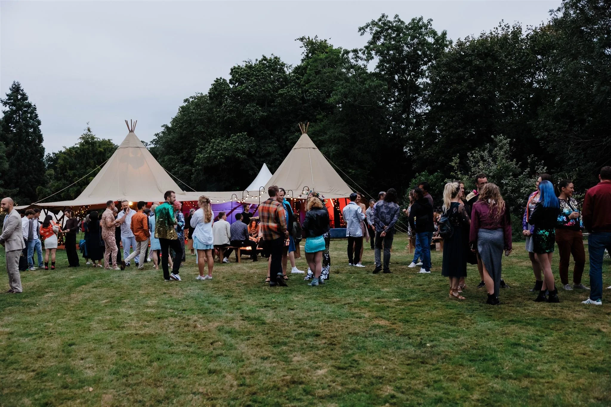 People gathering outdoors near two large beige tipis with festoon lights during an evening event.