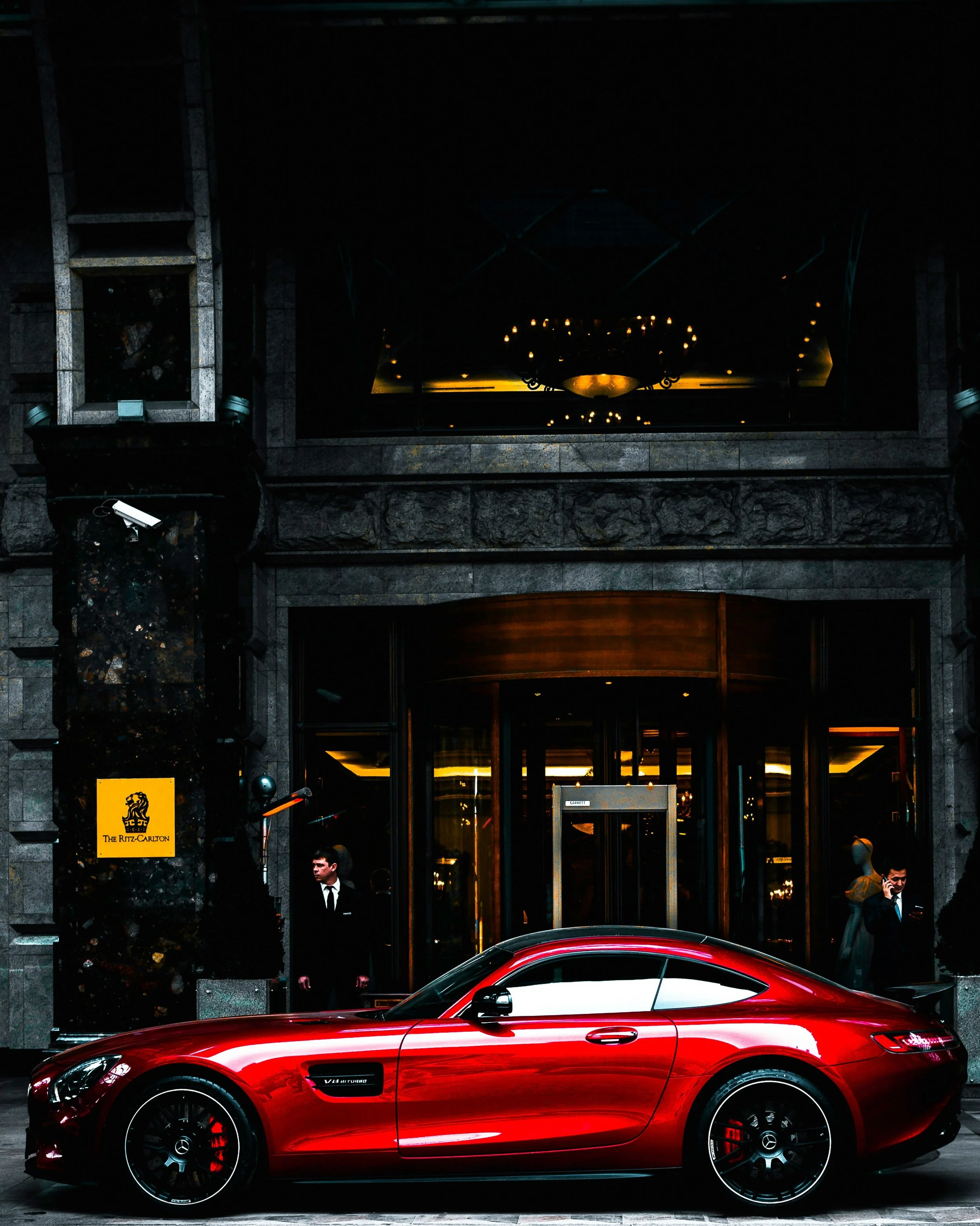 A red Mercedes-Benz sports car parked in front of The Ritz-Carlton hotel entrance, with hotel staff visible near the door and two people on the phone.