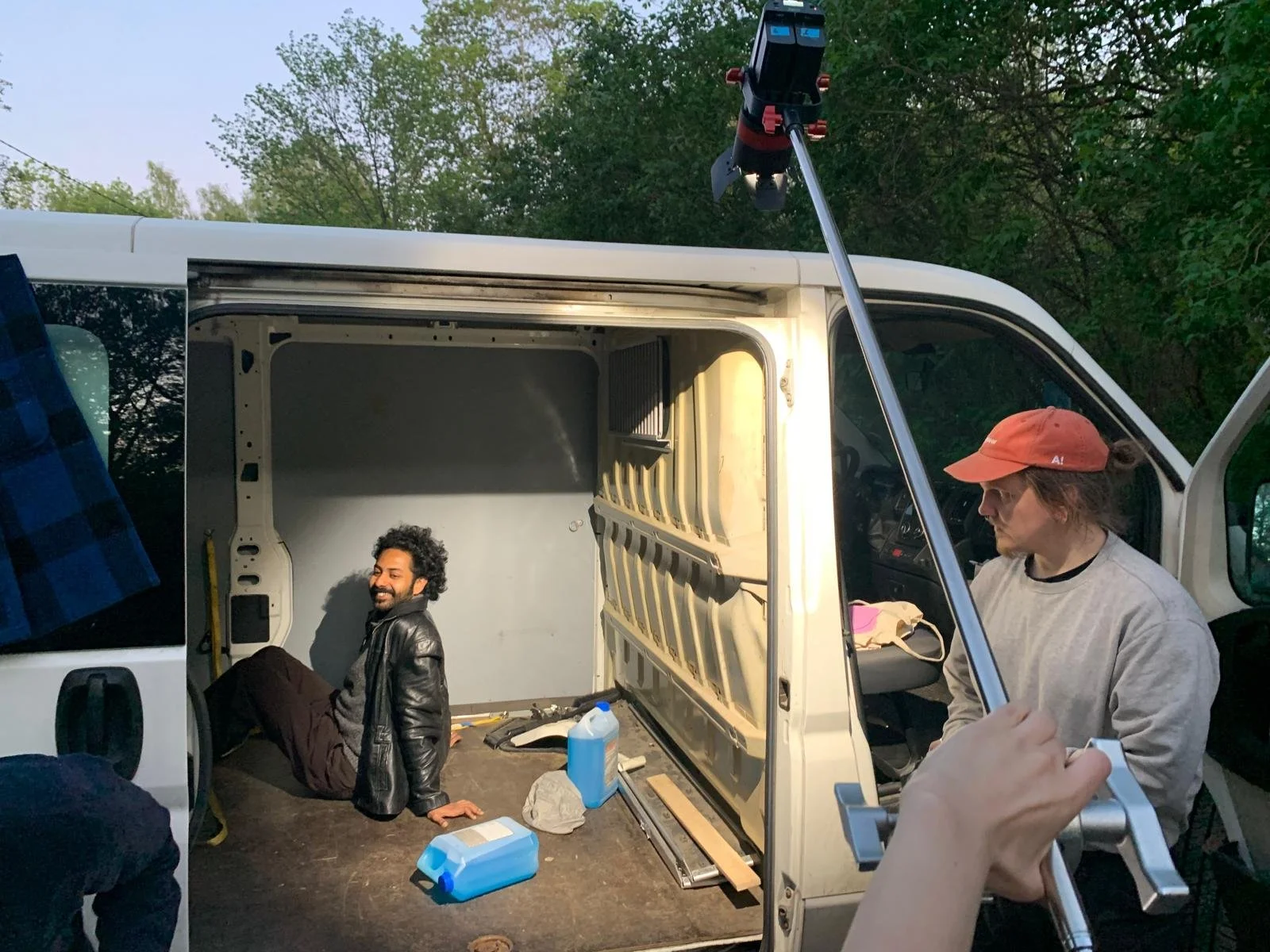 Two people working on a van outdoors with the door open. One person sitting inside the van smiling, and another person standing outside the van wearing a red cap. Staubwelt - BTS Poorman's process