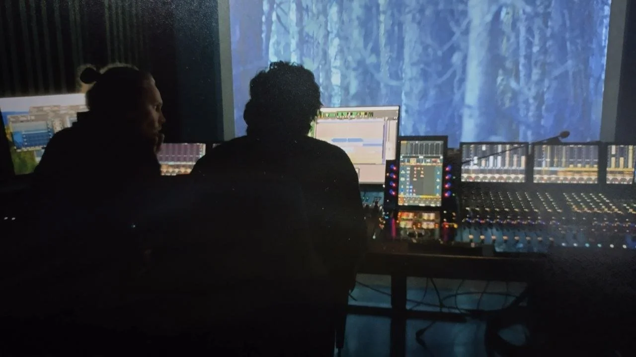 Two people working at a music production studio with multiple computer monitors, mixing consoles, and a backdrop of a snowy forest.