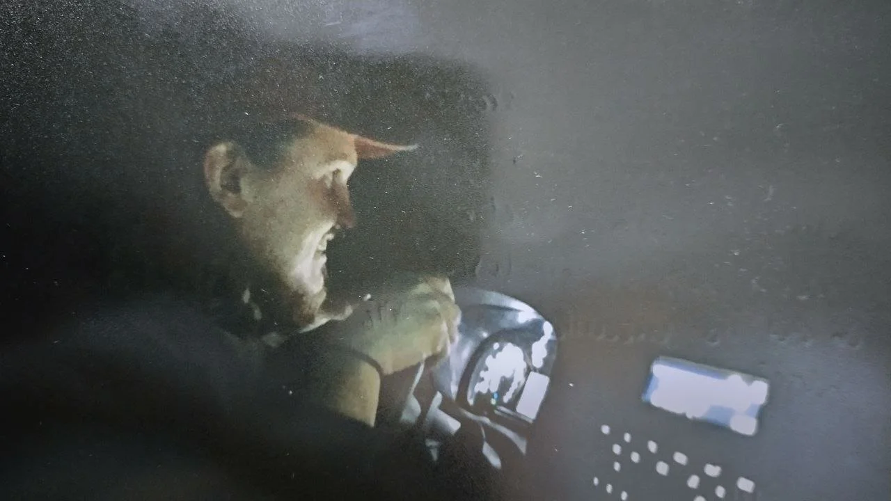 A man in a hat sitting in a vehicle at night, looking at a GPS device or screen, with rain on the window.