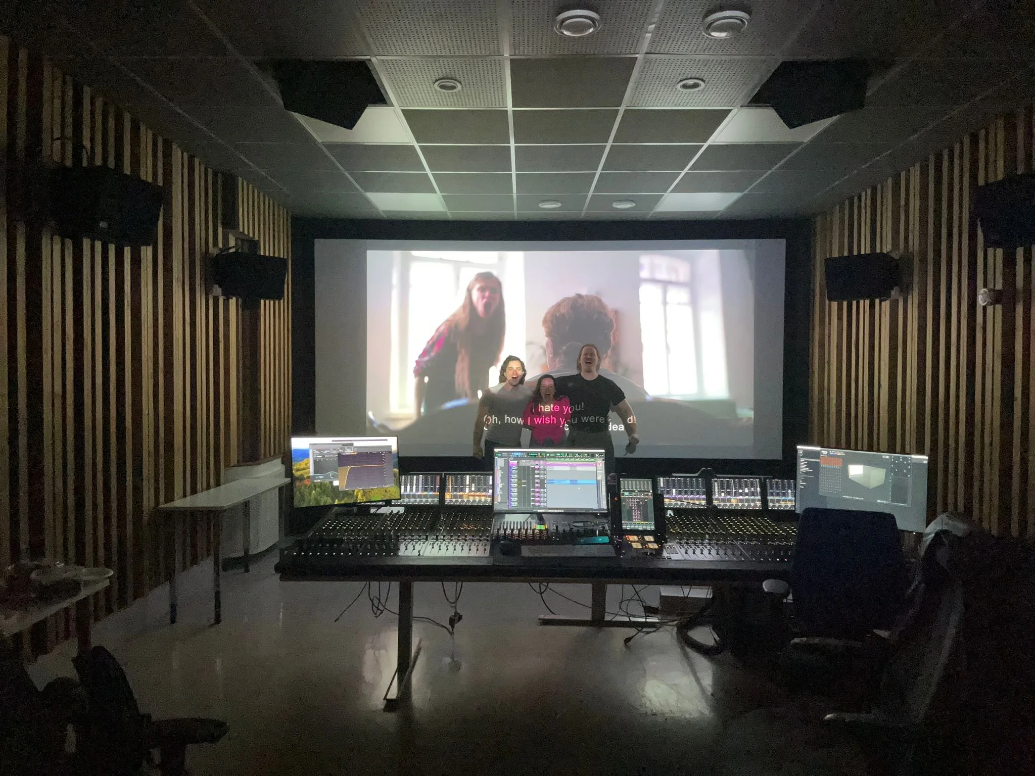 Recording studio with a large screen projecting a video of people, three of whom are visible laughing or talking. The studio contains multiple computer screens and a mixing console, with wooden panel walls and soundproofing.
