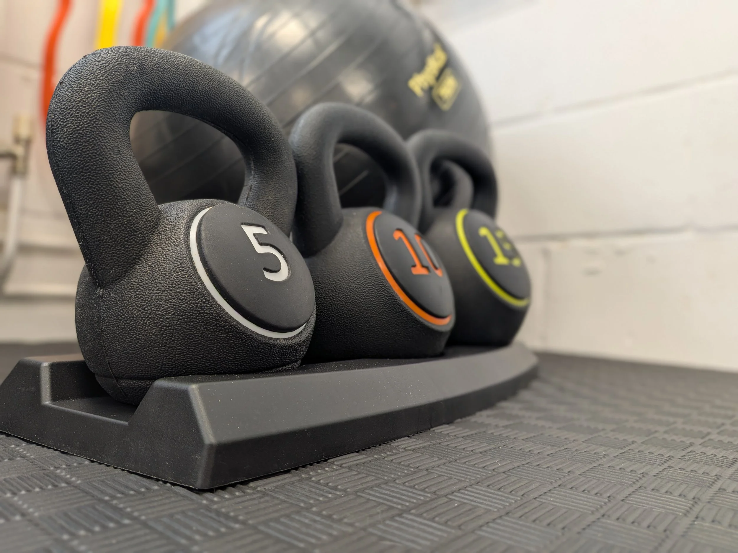 Three black kettlebells with colored number markings (5, 10, 12) placed on a black rubber workout mat against a white wall background