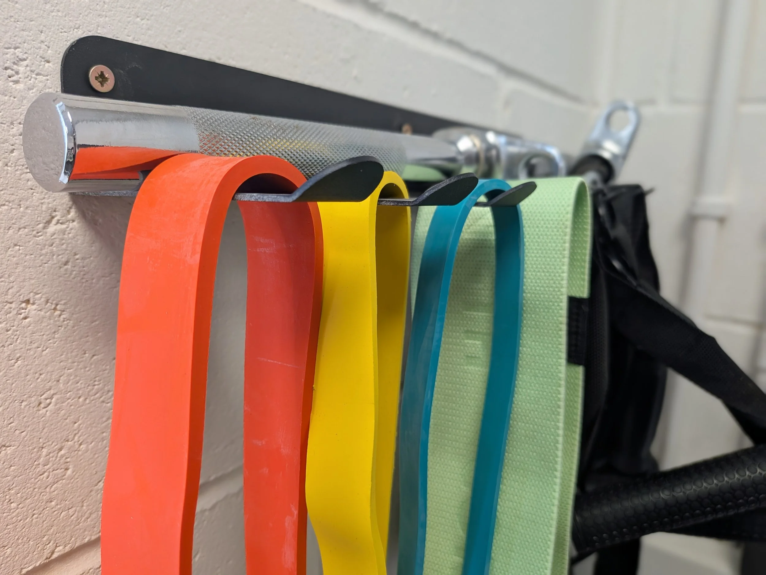 Colorful resistance bands hanging on a metal bar mounted on a wall, with bags visible in the background.