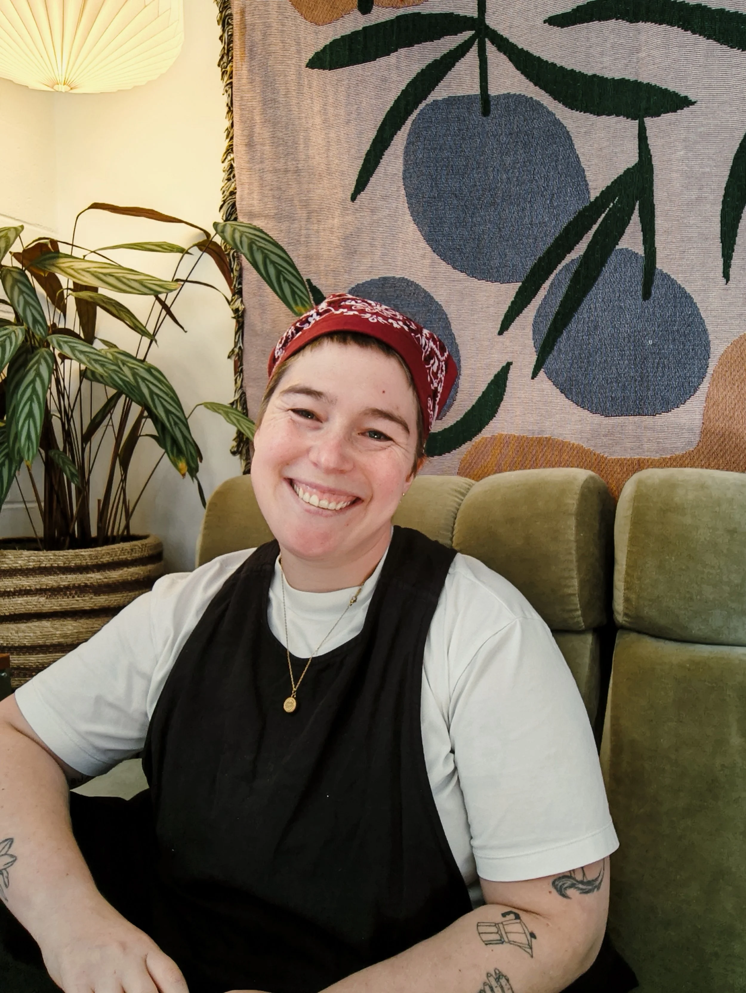 White person with short brown hair smiling, wearing a red bandana, black vest over a white T-shirt, sitting on a green couch in a room with indoor plants, a textile wall hanging with a stylized fruit pattern, and a lamp in the background.