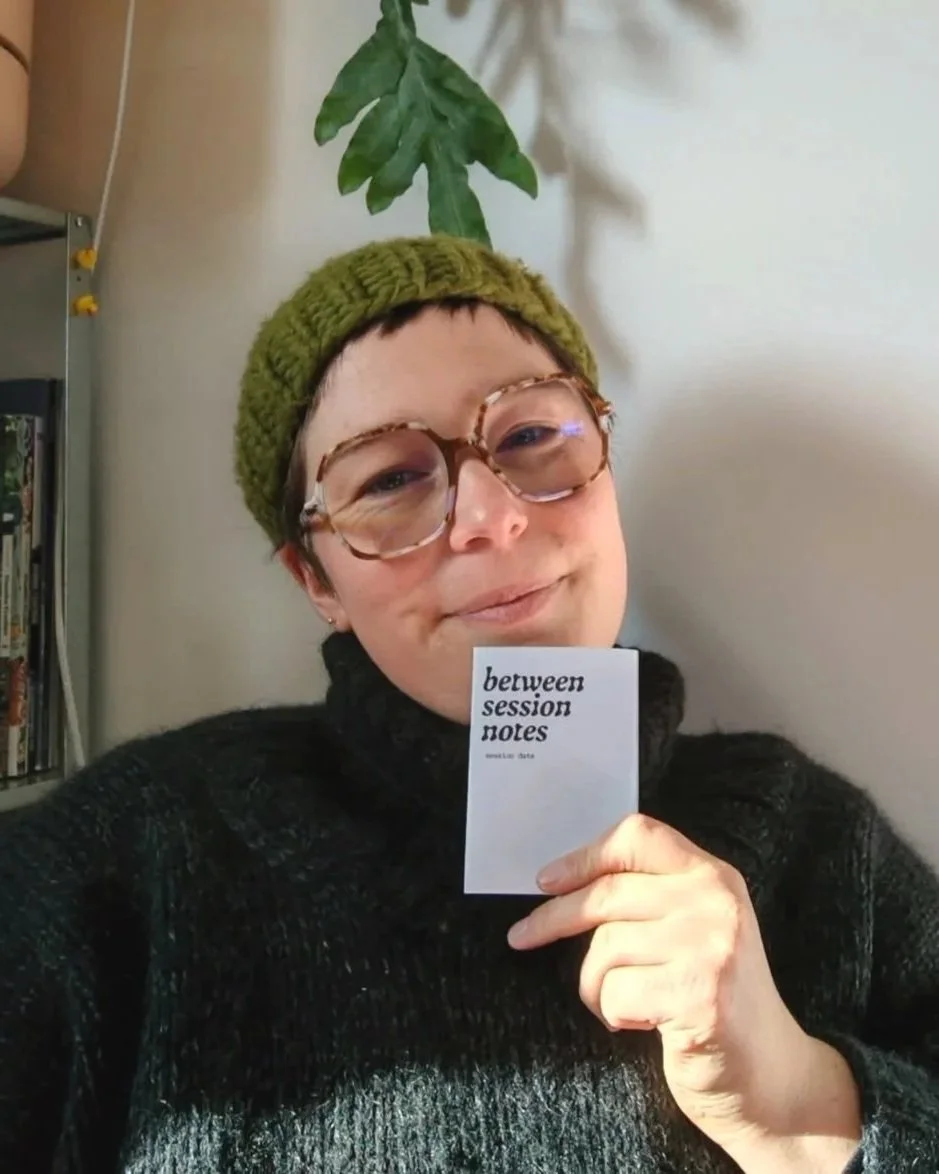 Rhiannon, a white person in a green beanie and big glasses, holds a mini-zine. The cover says "between session notes"