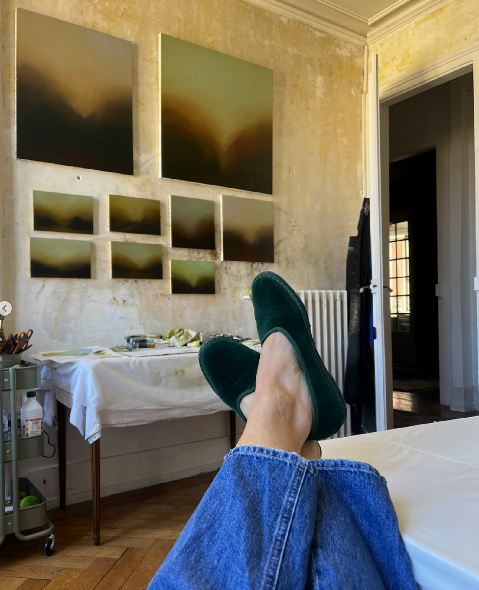 Personne allongée sur une table avec les jambes croisés, portant des chaussures vertes, dans une pièce avec des tableaux abstraits au mur.