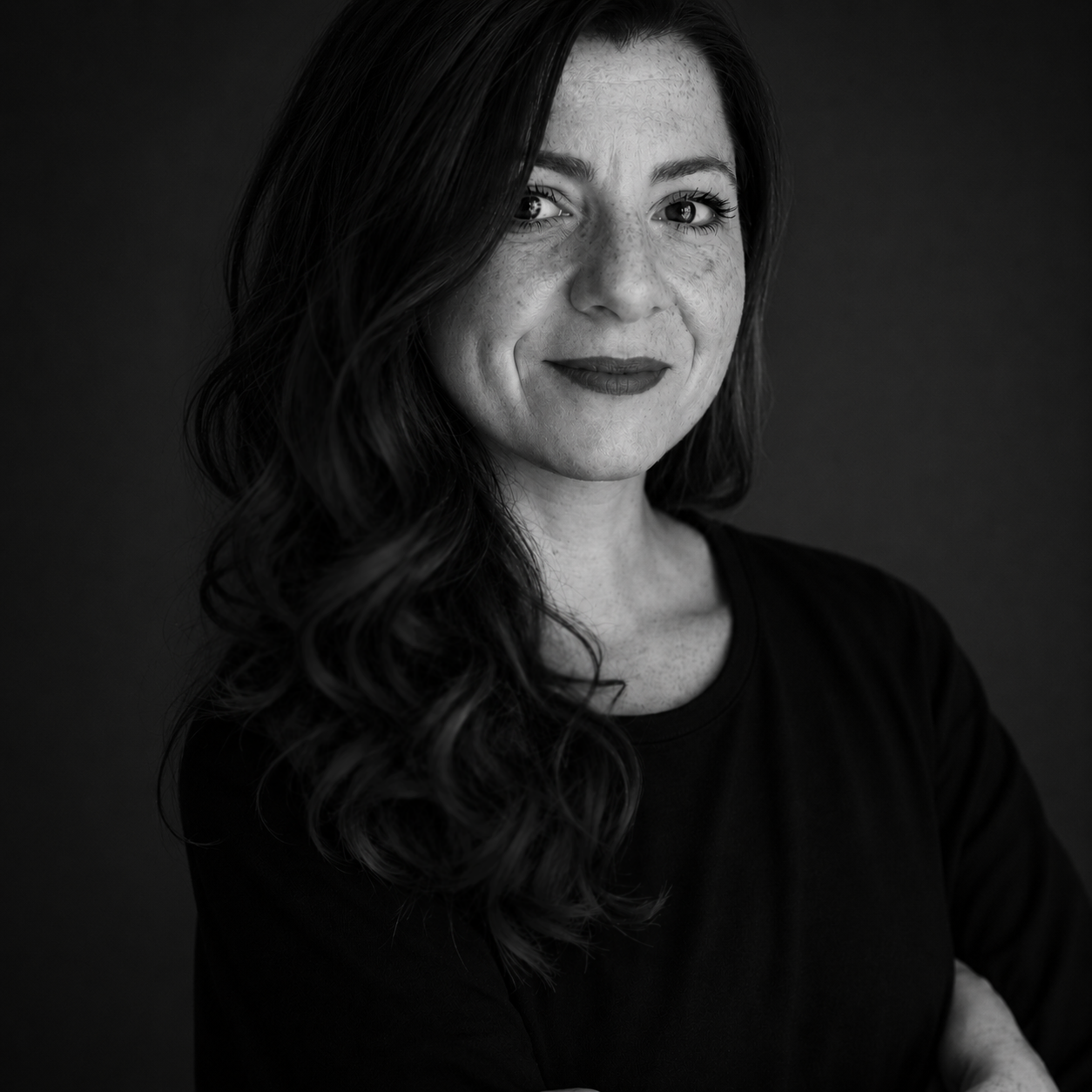 Portrait d'une femme avec des cheveux longs et bouclés, souriante, en noir et blanc, portant un haut noir, fond noir.