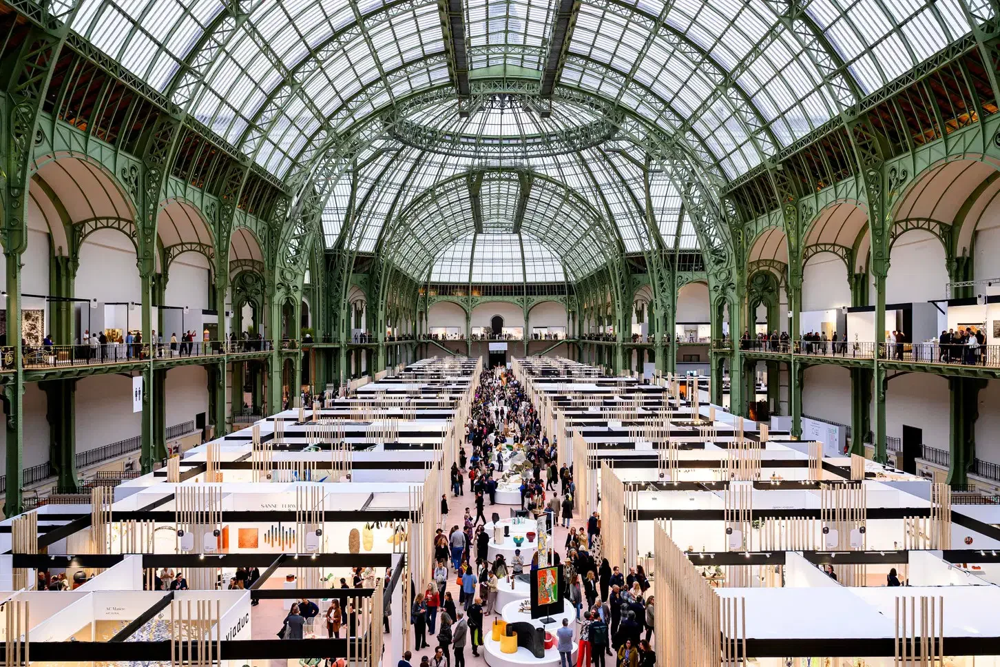 Vue intérieure d'un ancien bâtiment en verre et en fer transformé en galerie d'art ou marché avec plusieurs stands et nombreuses personnes.
