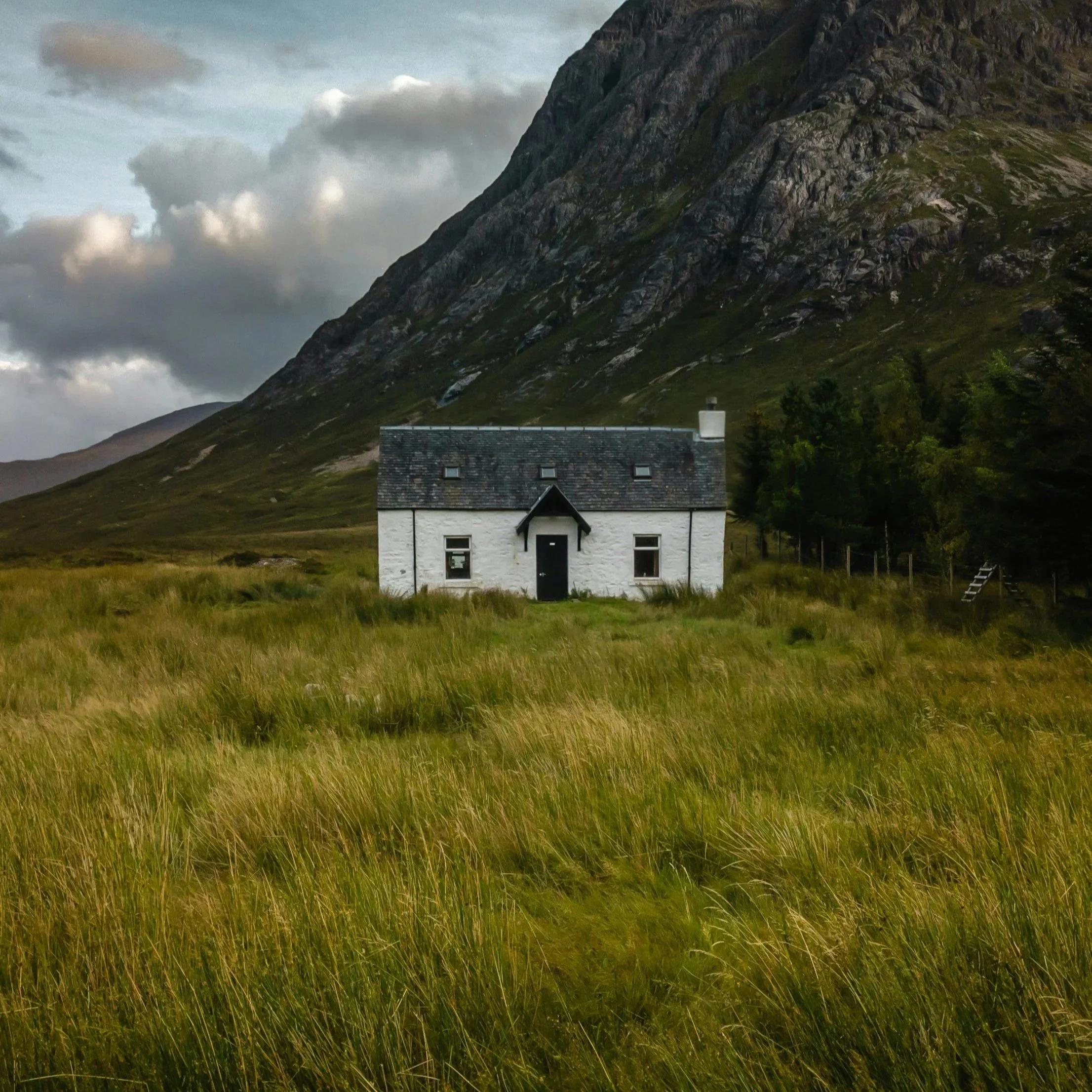 Vue d'une maison blanche avec une toiture en ardoise noire, située dans un paysage de campagne entouré d'herbes vertes, avec une grande montagne en arrière-plan et un ciel nuageux.