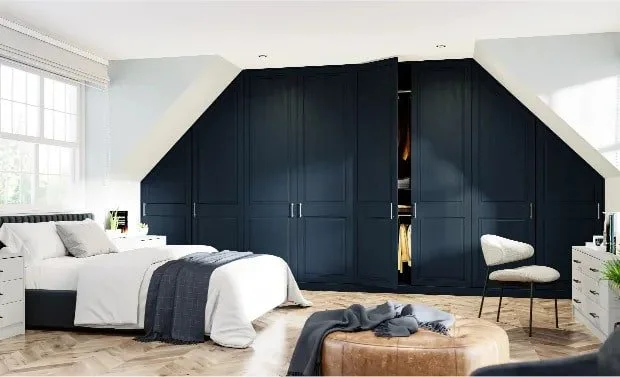 Modern bedroom with a white bed, dark blue built-in wardrobes, a cushioned ottoman, a beige chair, a nightstand, and large windows with blinds.