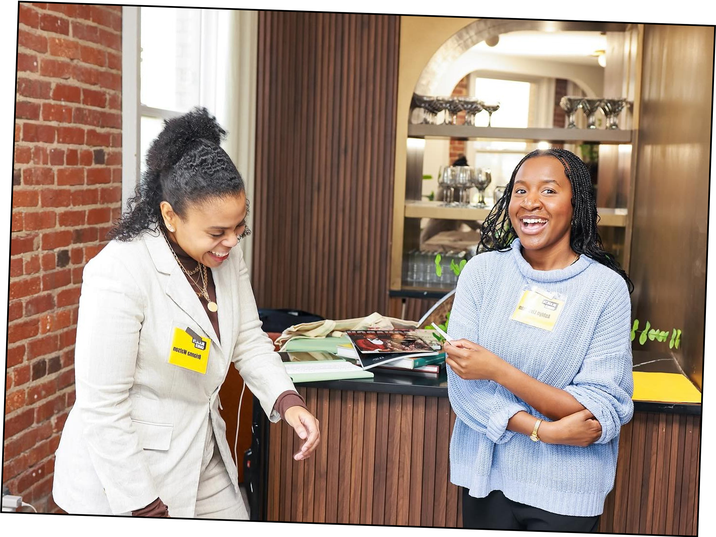 Two women are laughing and talking at a reception desk. One woman is wearing a white blazer with a yellow name tag, and the other is in a light blue sweater, also with a yellow name tag. They are in a well-lit, modern space with brick wall and wooden accents.