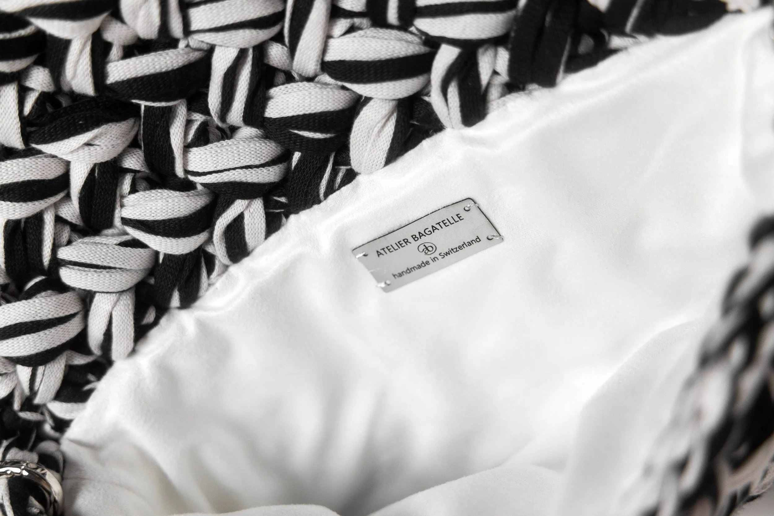 Close-up of a black and white striped woven bag with a white fabric interior label that reads 'ATELIER BAGATELLE' and 'handmade in Switzerland.'