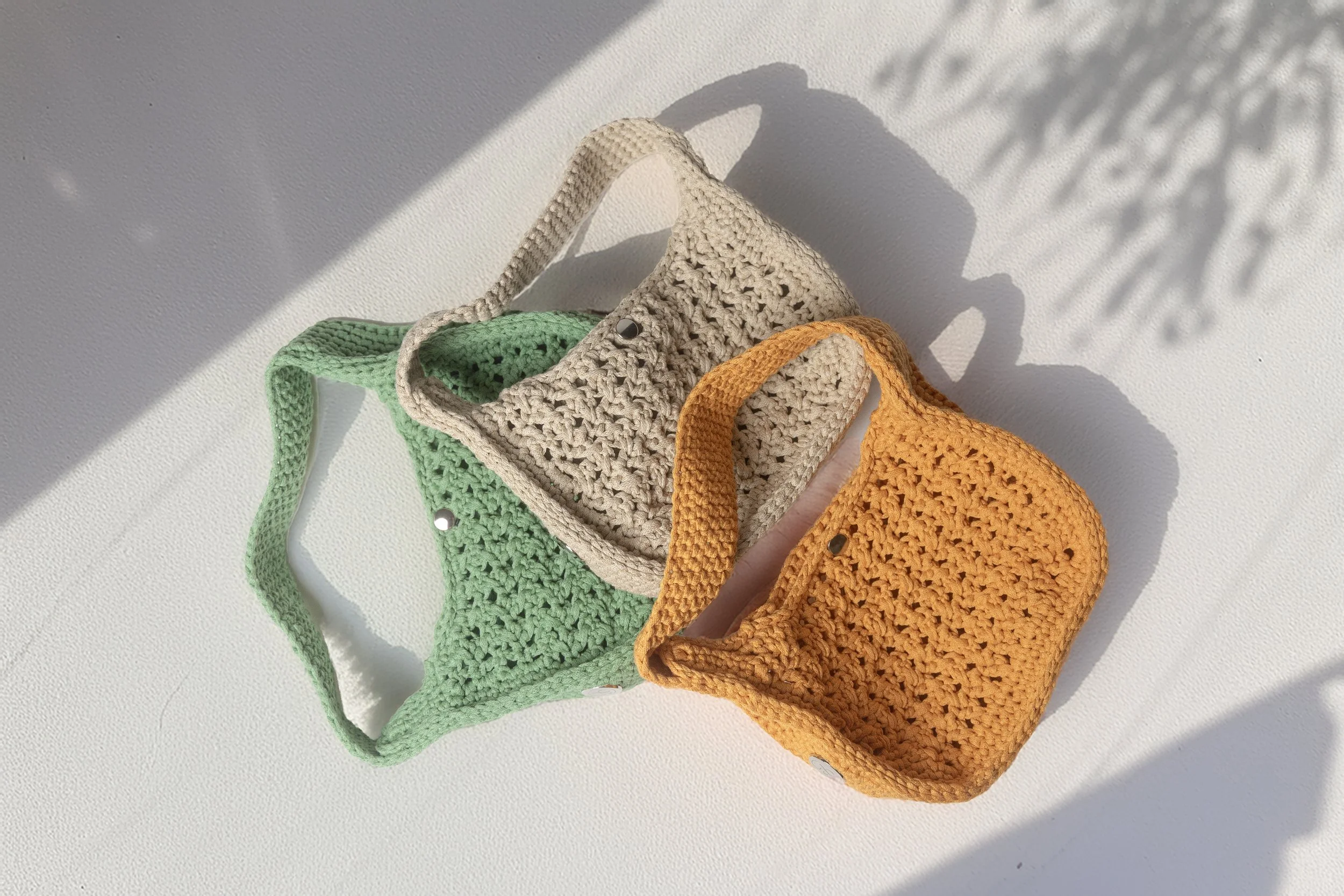 Three small crocheted bags in green, beige, and orange on a white surface casting shadows.