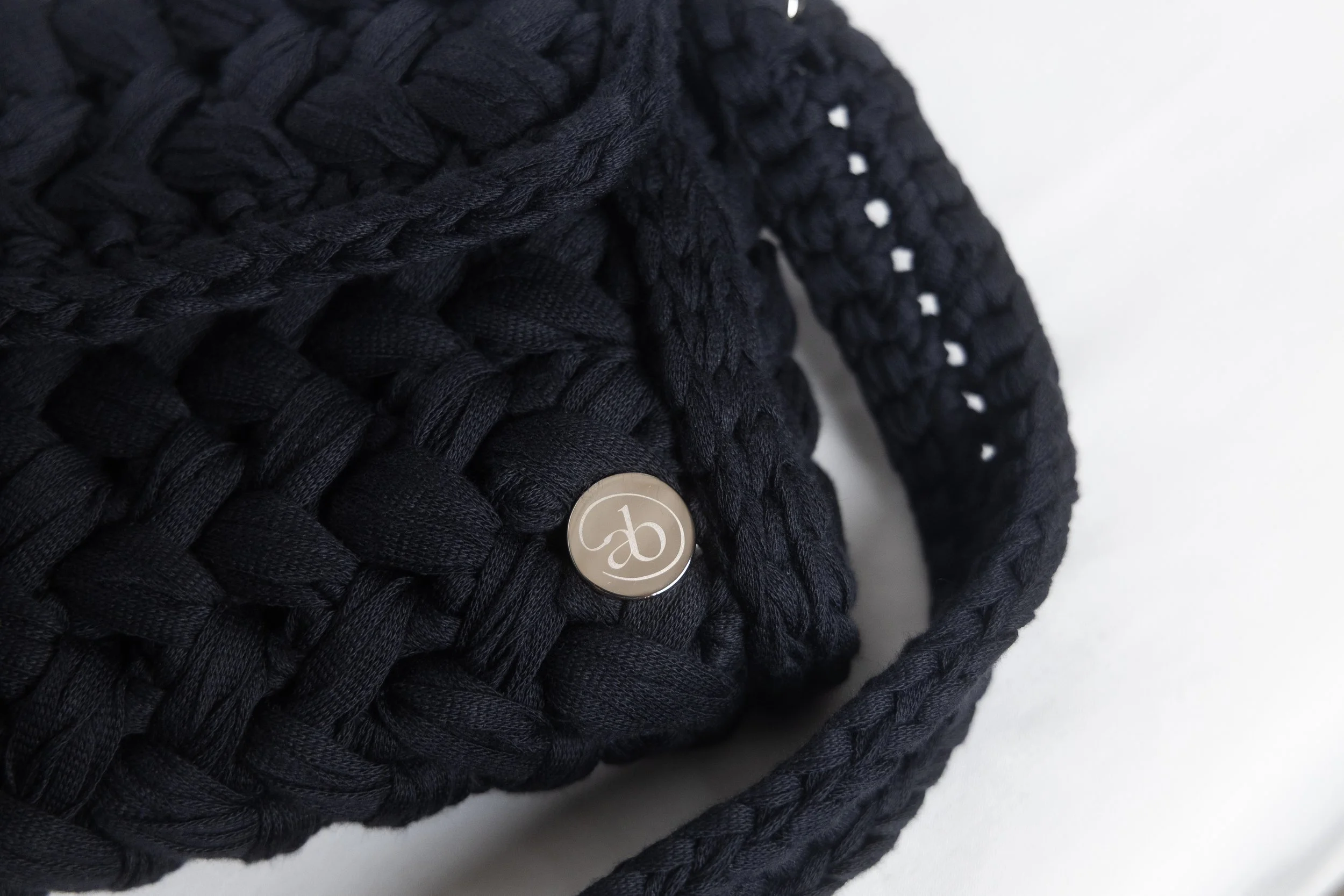 Close-up of a black braided knitted bag with a button featuring an embossed logo, on a white background.