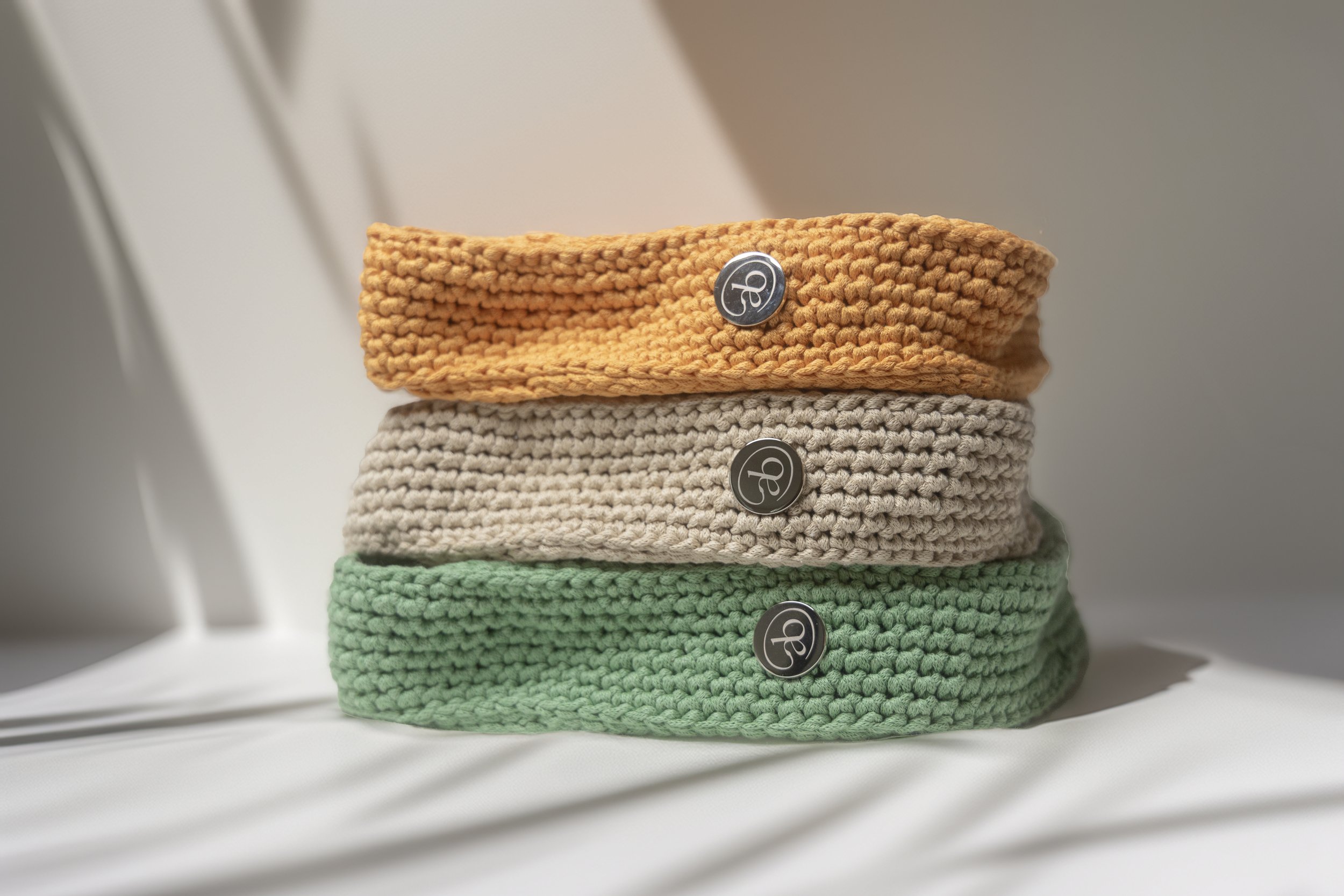 Stacked knitted headbands in orange, beige, and green with metallic logo buttons on each.
