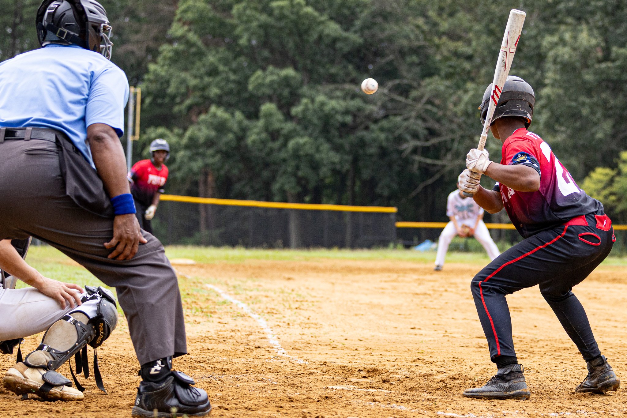 A baseball player in a red uniform preparing to hit a pitch at home plate, with an umpire crouching behind him and a player in the outfield in the background.