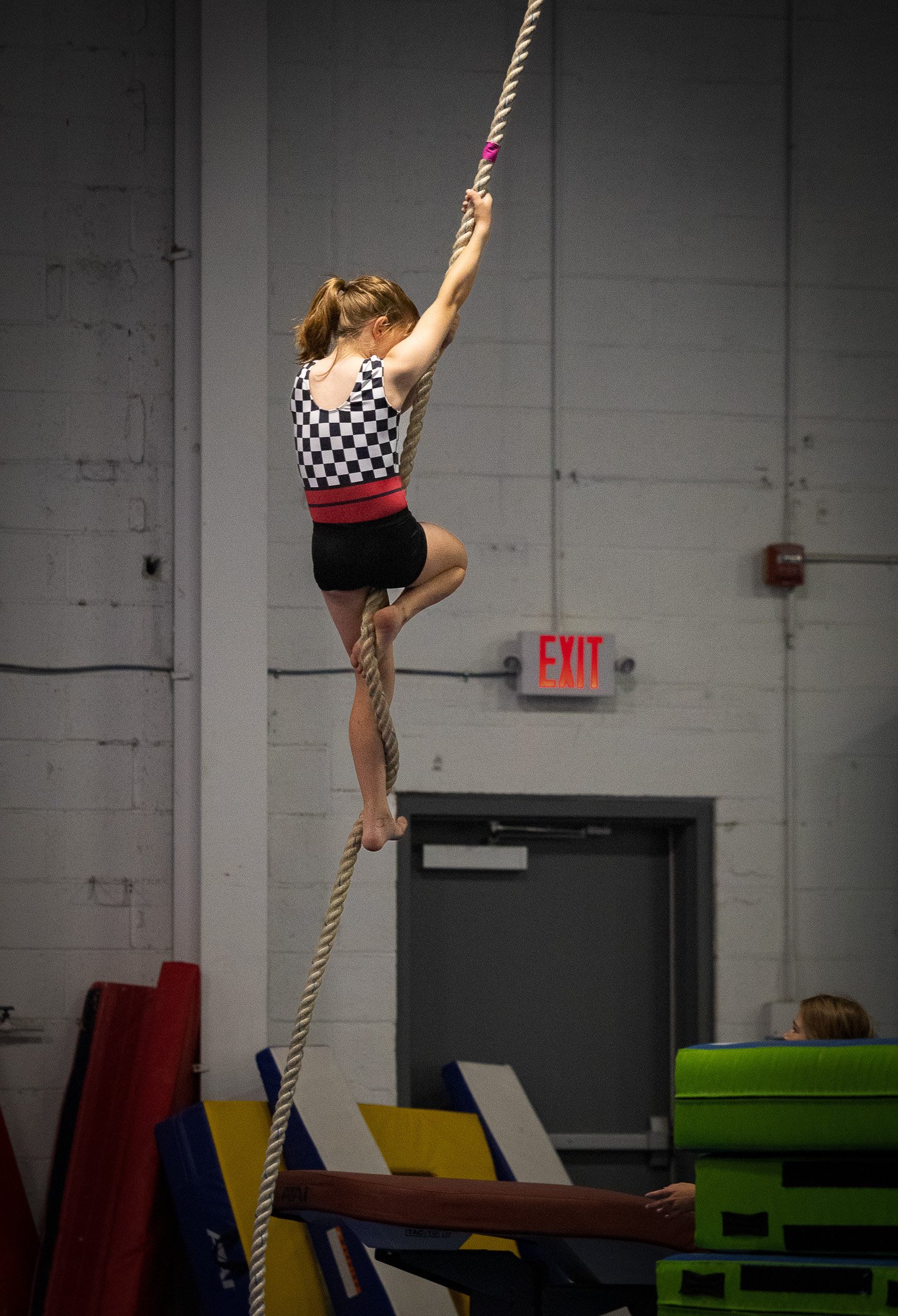 A young girl in a checkered top and black shorts climbing a thick rope in an indoor gymnasium, with an emergency exit sign visible in the background.