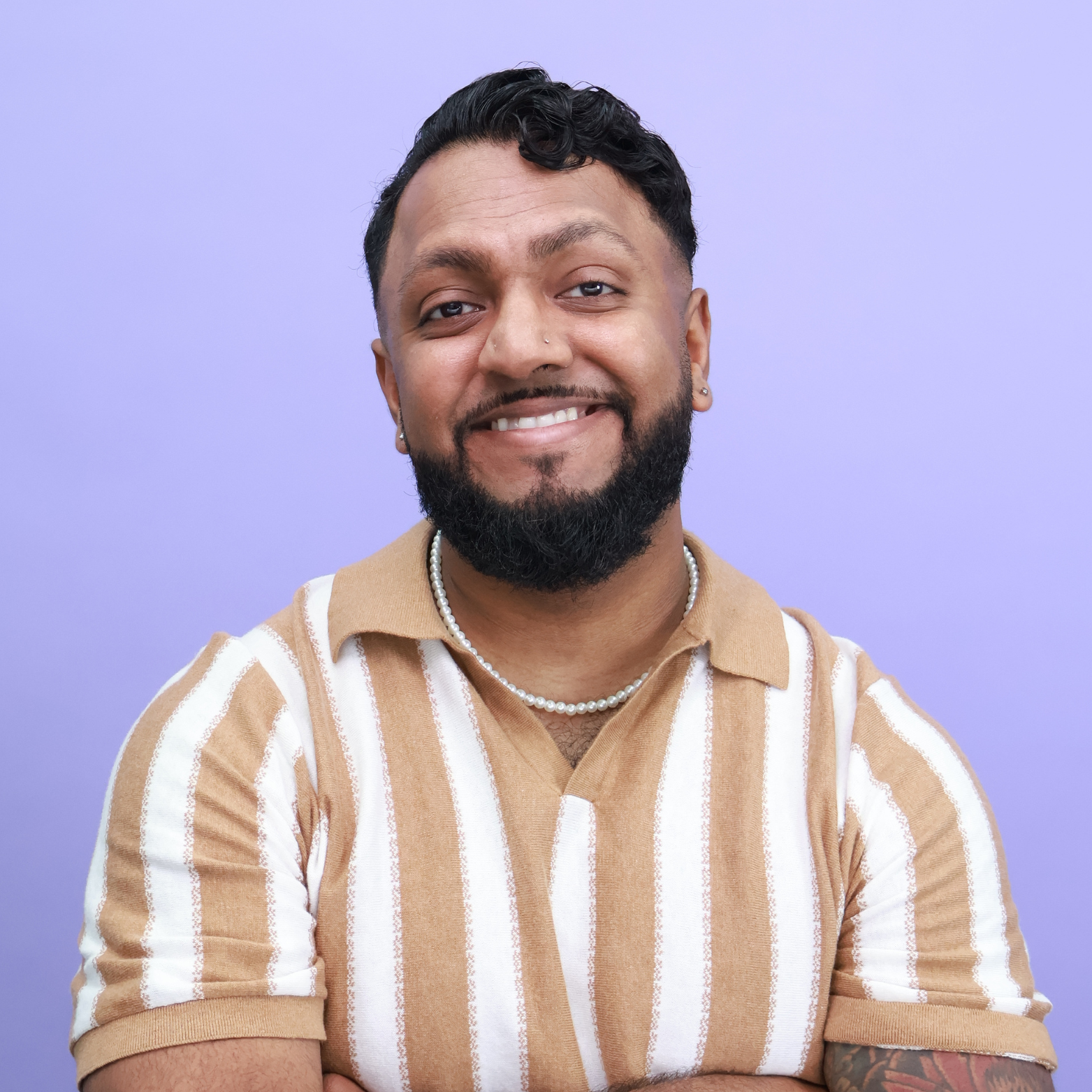 Portrait of a smiling man with a beard and mustache, wearing a tan and white striped polo shirt, pearl necklace, and earrings, standing against a purple background.