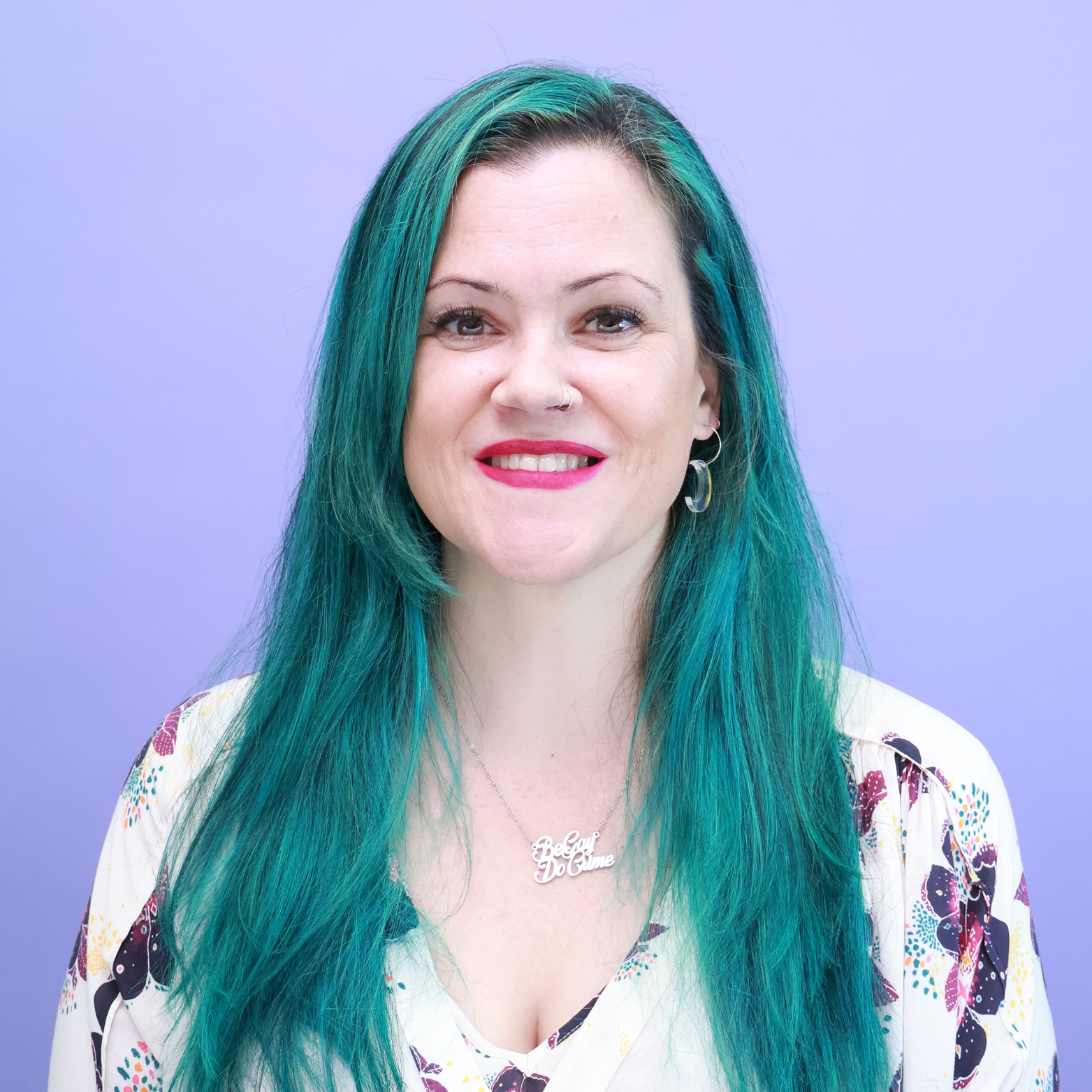 A woman with long, teal hair smiling in front of a solid purple background, wearing a white floral blouse, earrings, and a necklace that says 'Believe in Yourself'.