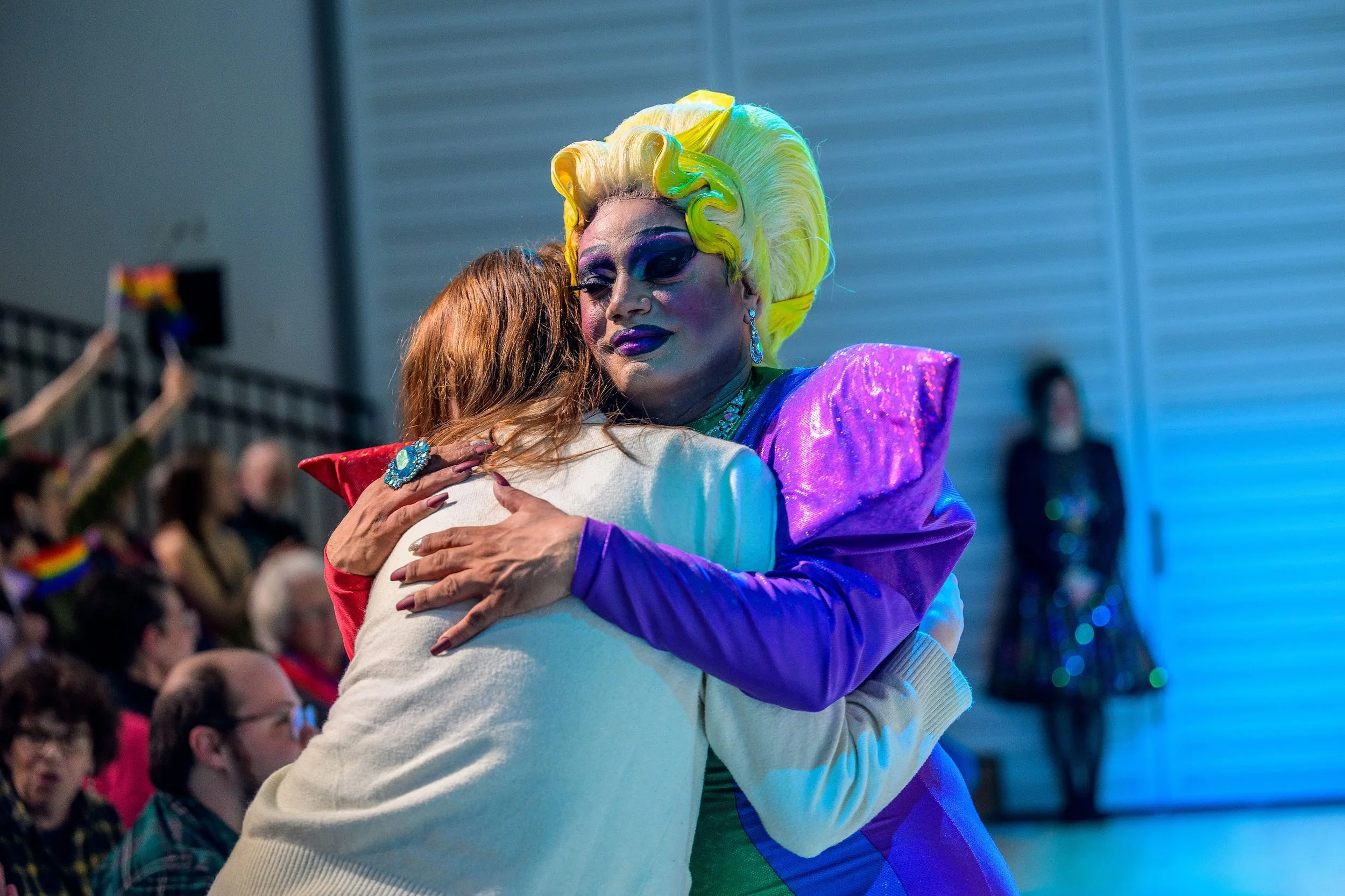 Two women embrace in a hug at a public event, one dressed in colorful, glamorous clothing with bright yellow hair, large earrings, and dramatic makeup, while the other has red hair and a cream-colored outfit, with a crowd of people and a person in the background wearing a dark, shiny outfit.