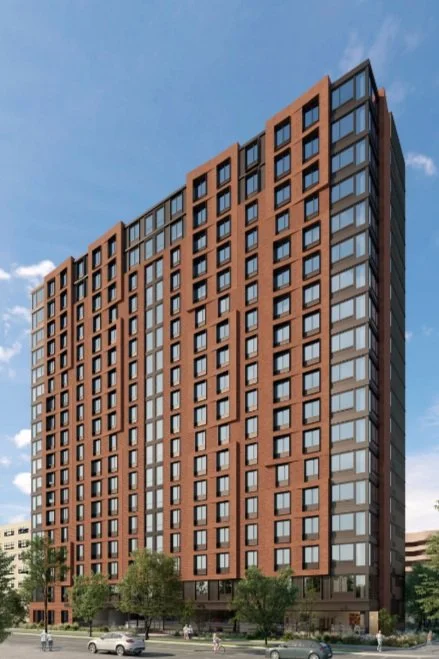 Tall modern apartment building with a brick facade and large windows, set against a blue sky.