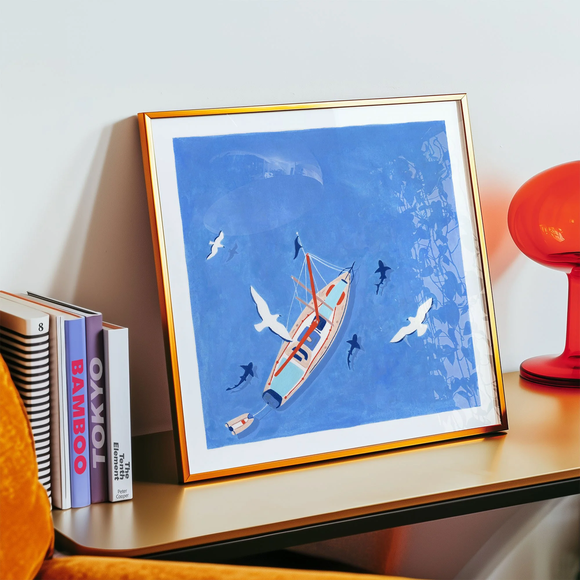 A framed picture of a sailboat on blue water surrounded by flying birds, leaning against a wall on a white shelf. Books are to the left, and a red decorative lamp is to the right.