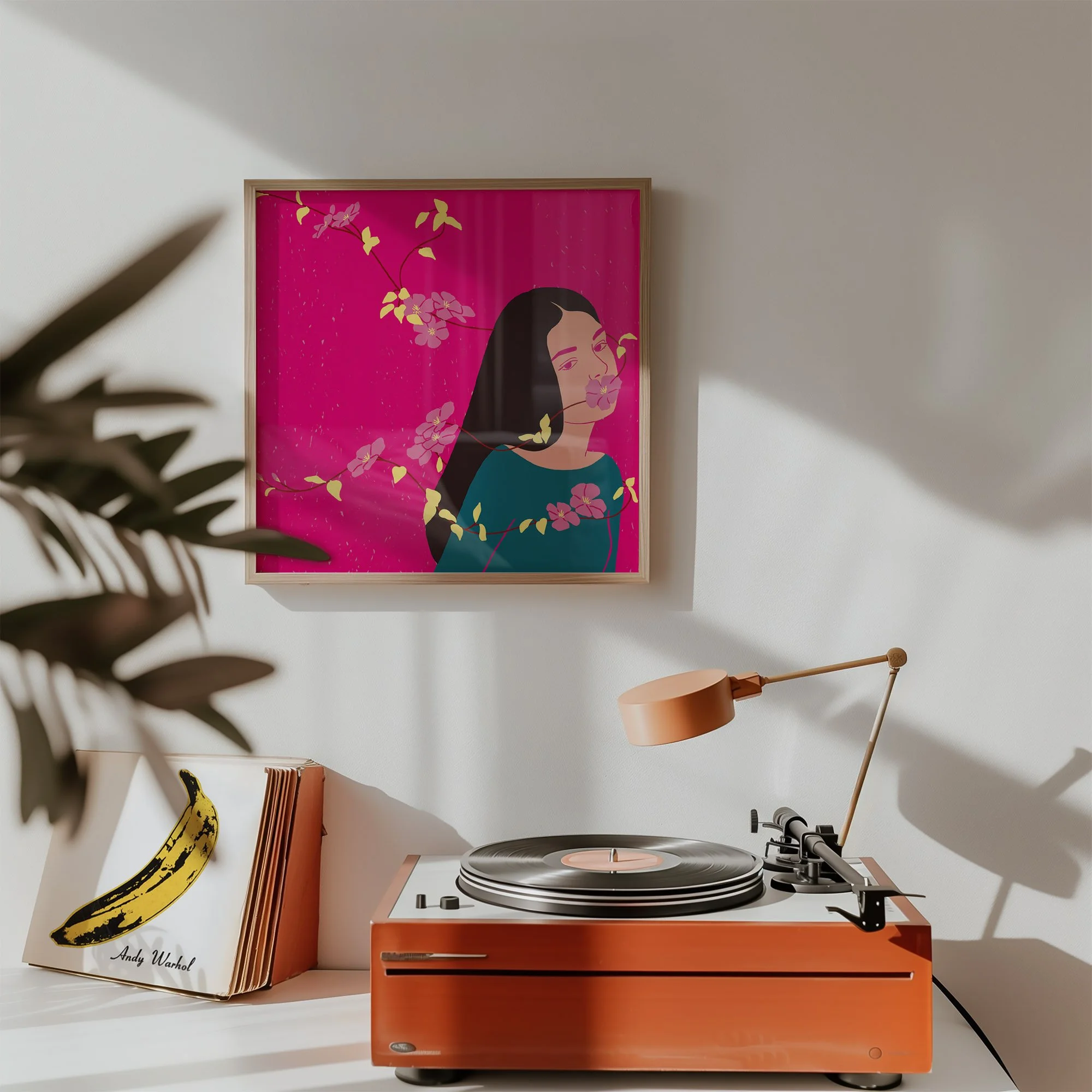 A vinyl record player on a white surface with a framed art piece of a woman with long black hair, wearing a dark top, with pink and yellow floral elements on a pink background, hanging on a white wall. A book with a banana illustration and the signat