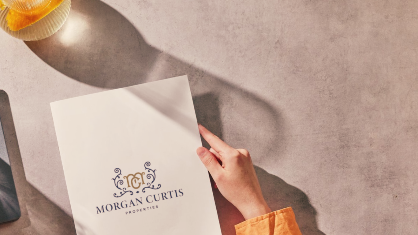 A person holding a white folder with the logo and name of Morgan Curtis Properties printed on it, on a beige surface with a yellow-orange object partially visible.